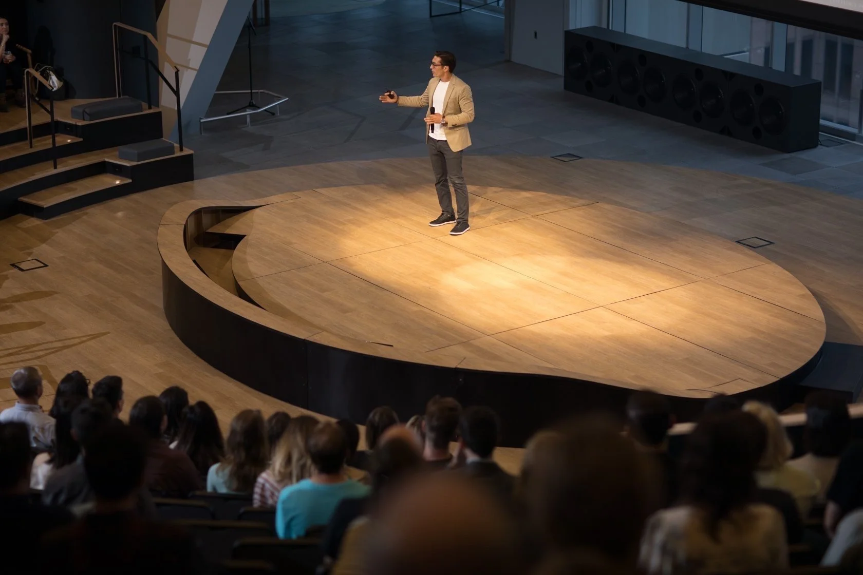 A man in a beige blazer giving a presentation on a stage with a wooden floor, audience seated watching him, in a modern auditorium.