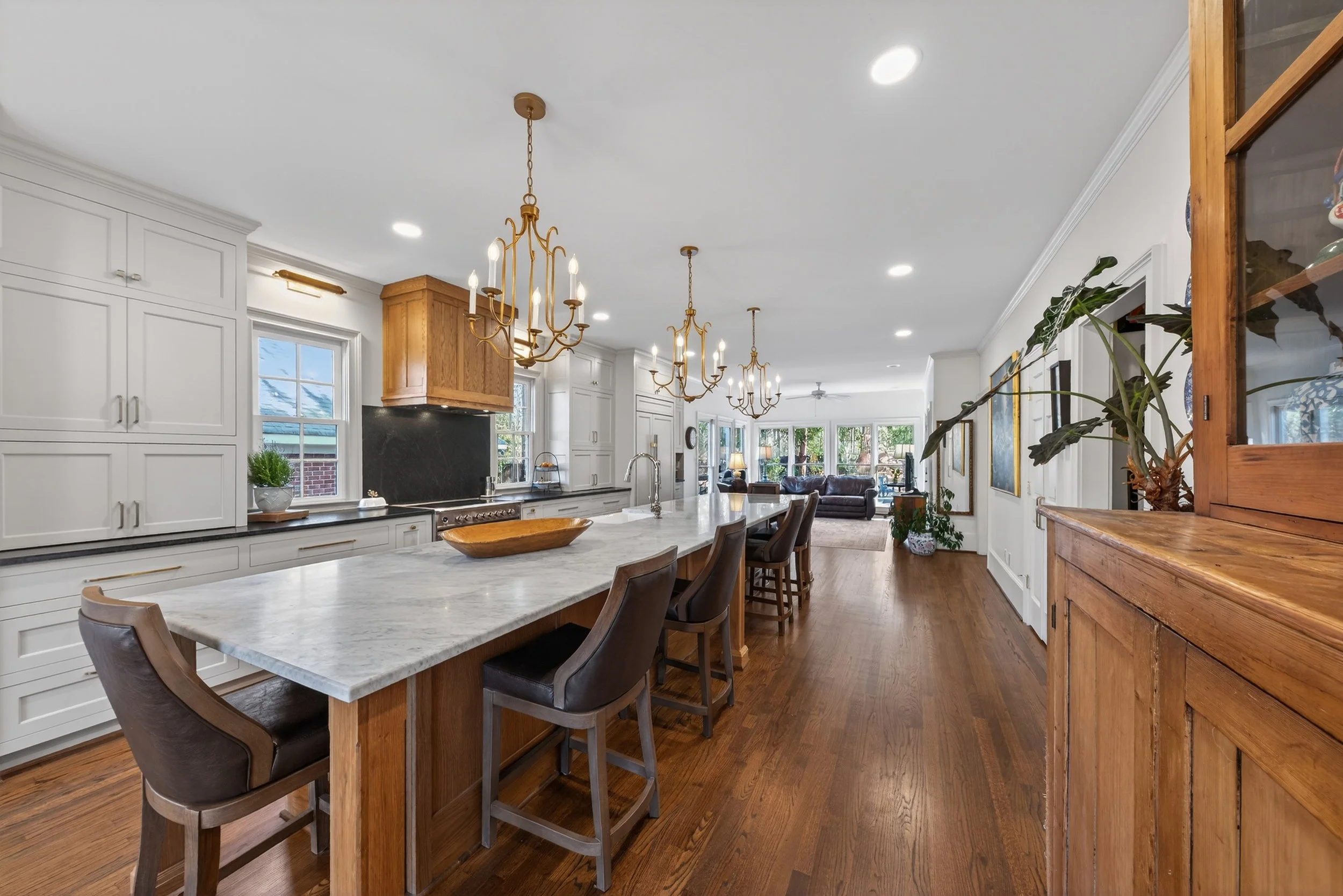 Open concept kitchen with white cabinets, a large marble island, wooden accents, gold chandeliers, and a view of the living room with sofas and bay windows.