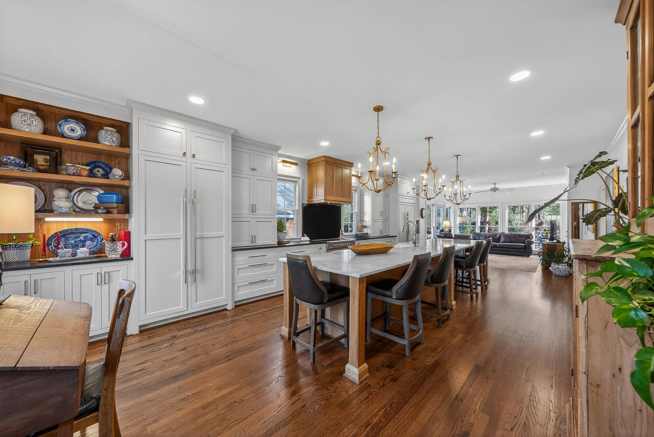 Open kitchen with white cabinetry, a central island with marble countertop, and wooden flooring. Gold chandeliers hang above the island. A breakfast bar has four black chairs. In the background, a living room with sofas, tables, and large windows.