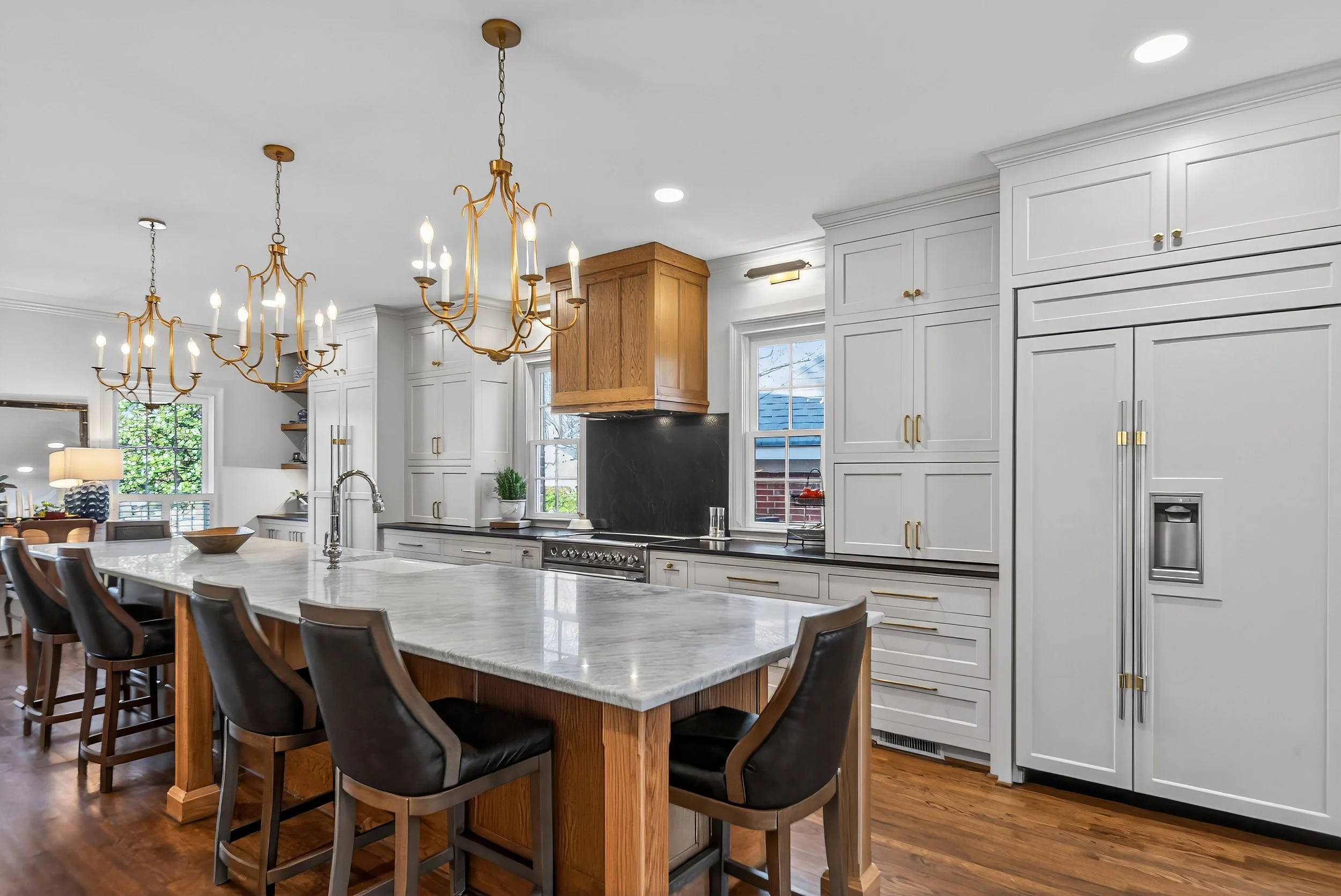 Modern kitchen with white cabinets, gold hardware, black countertops, a central island with a marble top, pendant chandeliers, and a window showing a roof outside.