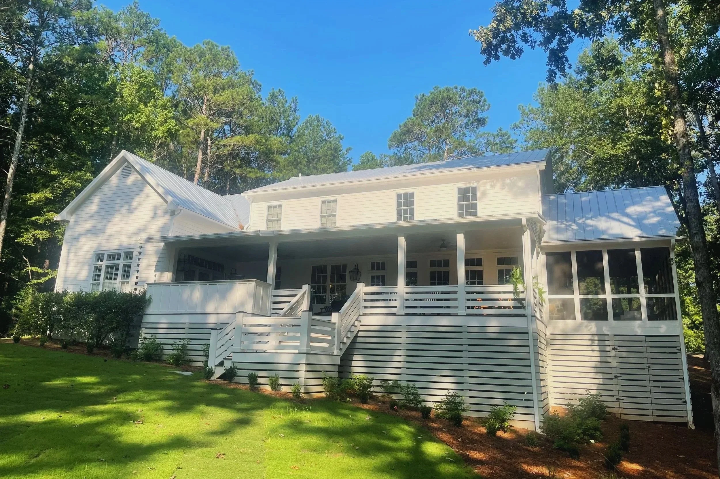 White two-story house with a screened-in porch, surrounded by trees and a well-maintained lawn, under a clear blue sky.
