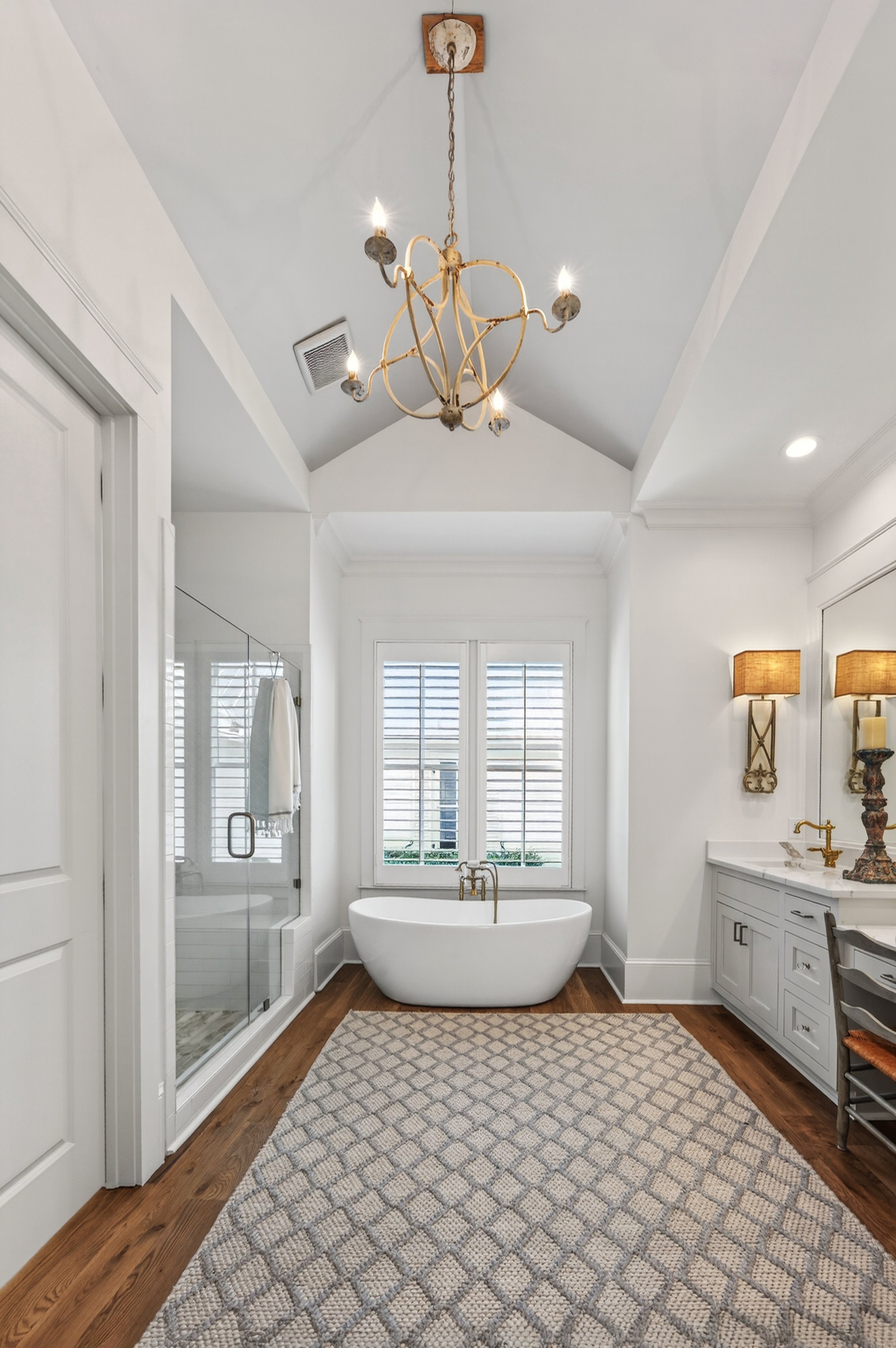 A luxurious bathroom with a white clawfoot bathtub, a glass-enclosed shower, hardwood floors, a large window with shutters, a white vanity with gold fixtures, and elegant wall sconces with lampshades. A chandelier hangs from the vaulted ceiling.