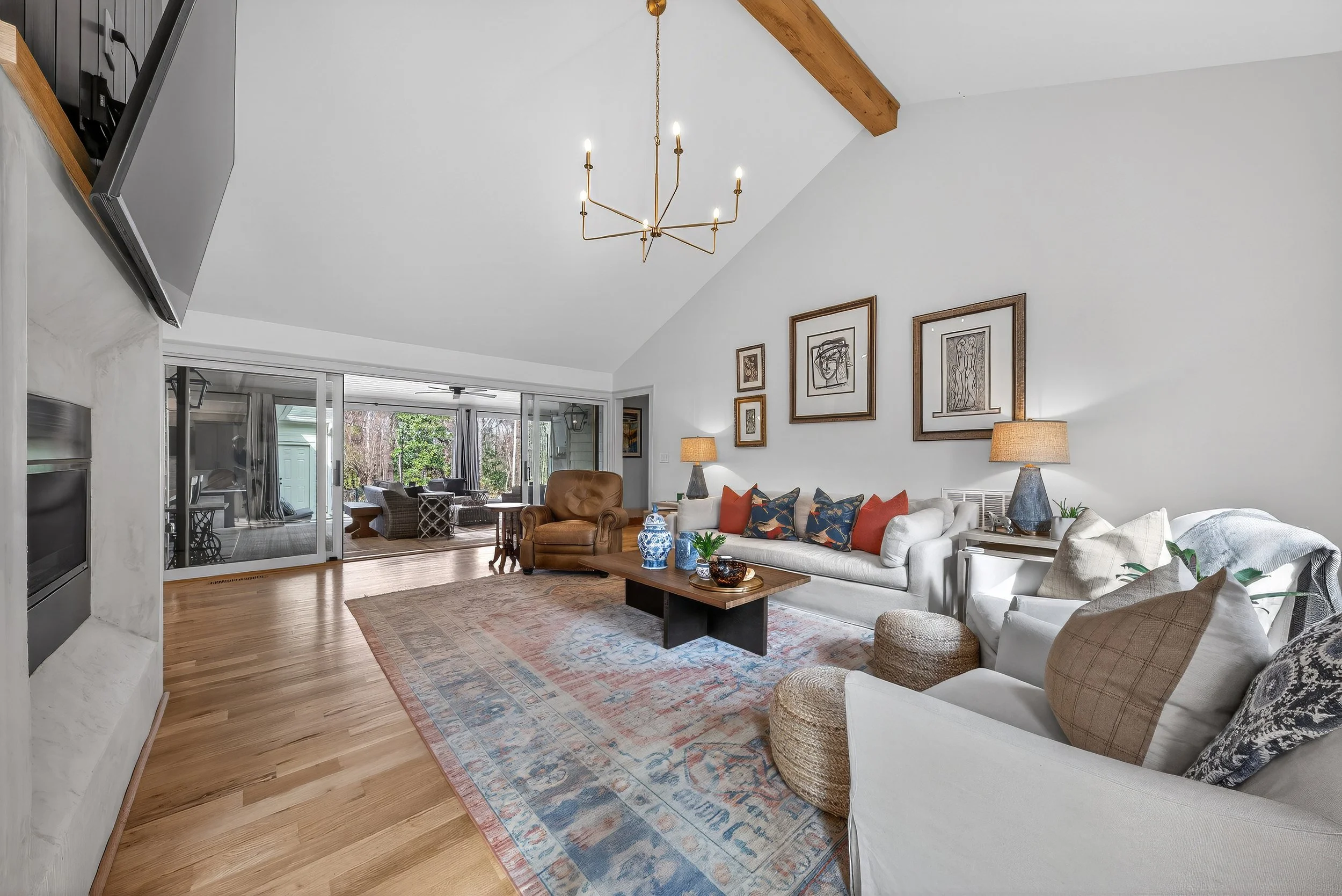Living room with a white sofa, colorful pillows, a brown leather armchair, wooden coffee table with decorative items, a patterned area rug, and wall art. There's an open doorway leading to a sunroom with outdoor furniture and trees visible outside.