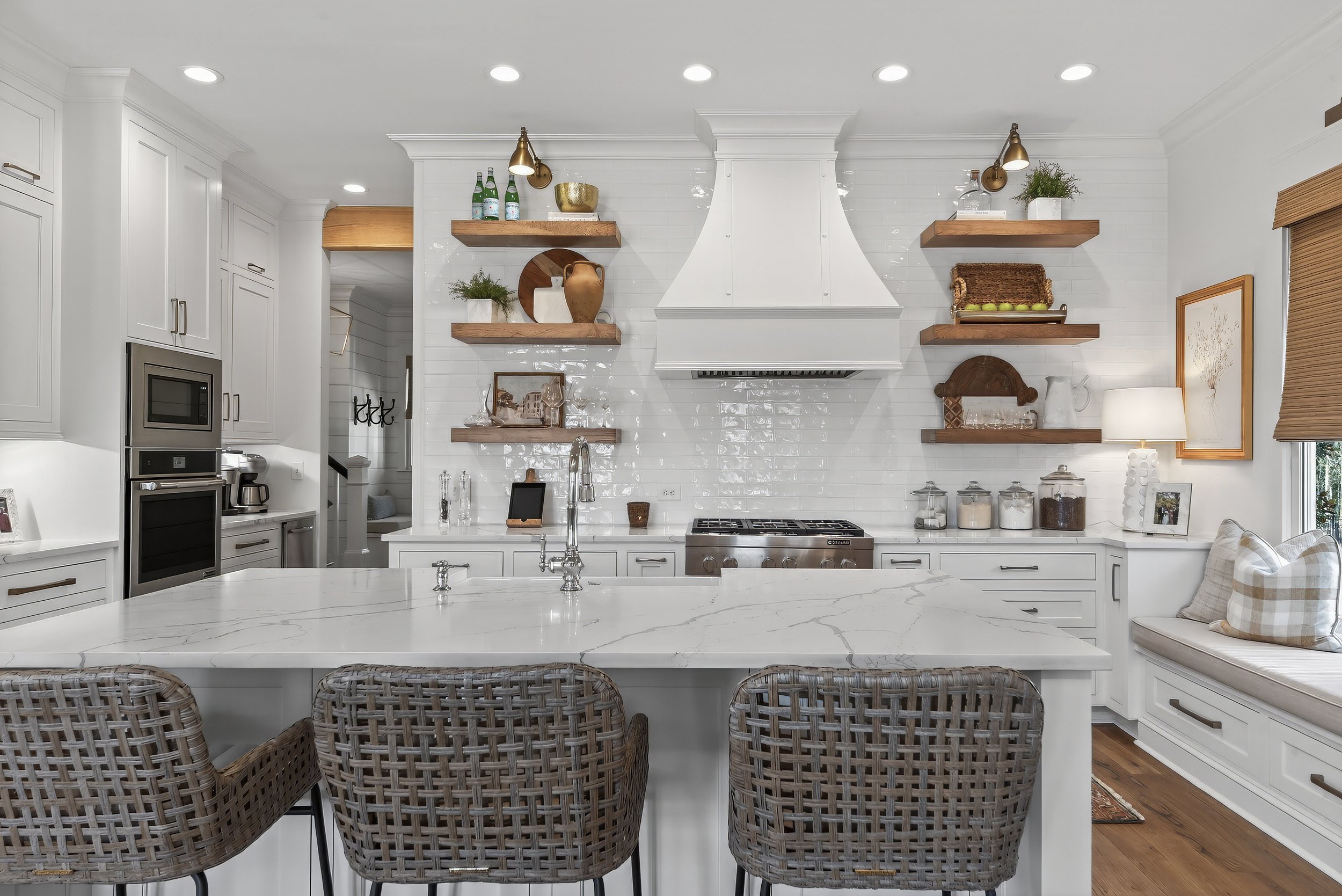 A bright, modern kitchen with white cabinets, a marble island with a built-in sink, open wooden shelves with decorative items and dishes, a window seat with cushions, and stainless steel appliances.