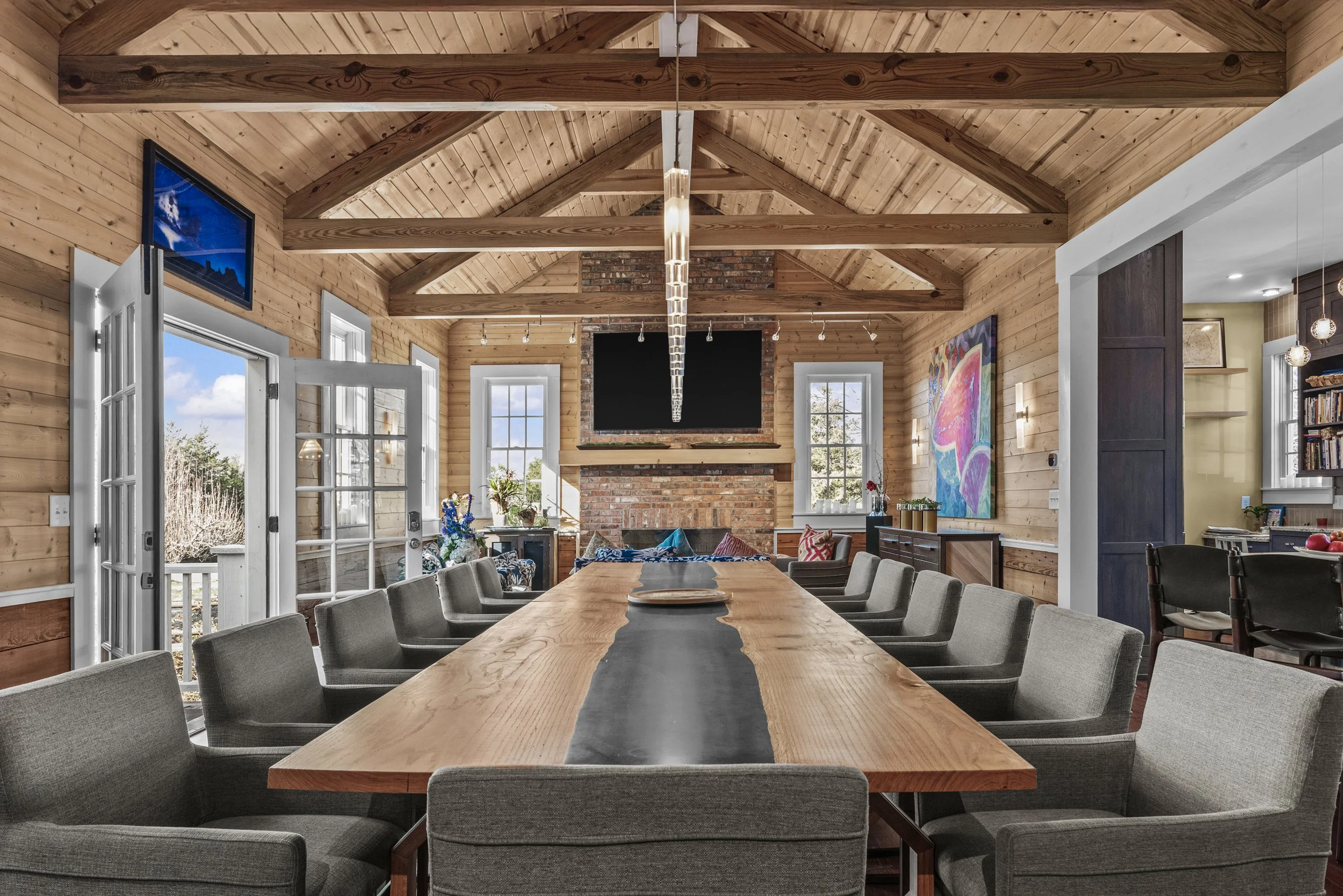 Spacious dining room with a large wooden table surrounded by gray upholstered chairs, wood-paneled walls, and a vaulted ceiling with exposed wooden beams. Features a brick fireplace, large windows, and colorful artwork on the wall.