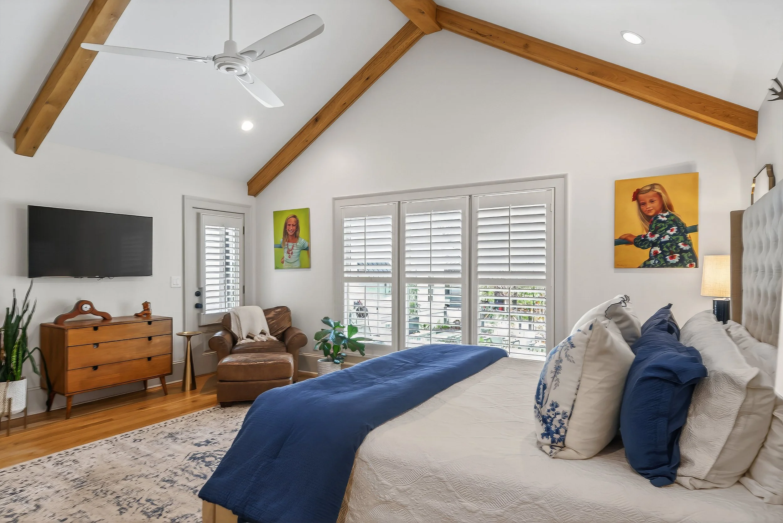 A bedroom with a vaulted ceiling, wooden beams, and a ceiling fan. There is a large bed with white and blue bedding, a wooden dresser with a flat-screen TV, and a brown armchair with a throw blanket. The room has three windows with white shutters, an