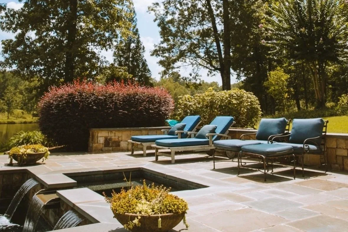 Outdoor patio with lounge chairs, potted plants, a small waterfall, and a stone wall, overlooking a yard with trees and a lake.