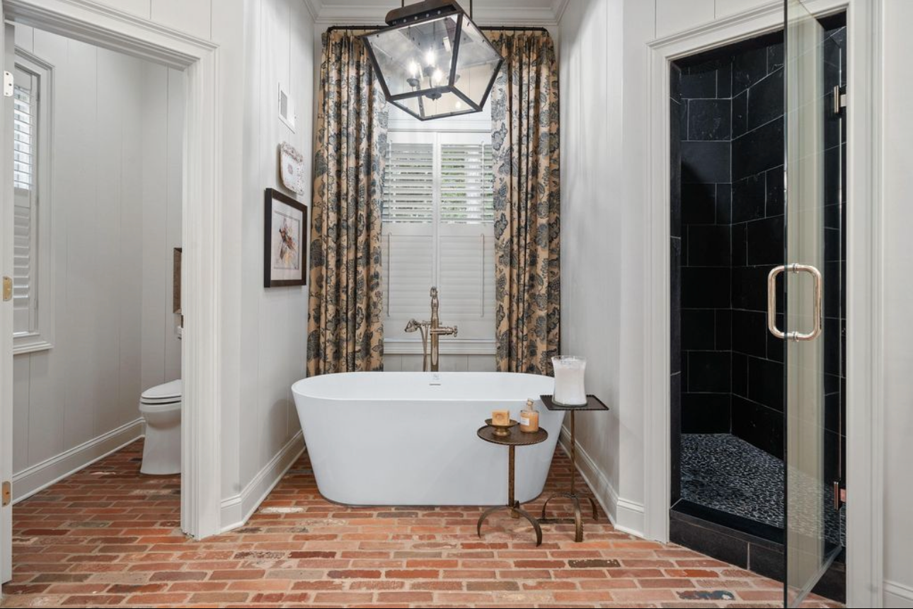 Bathroom with brick floor, white walls, a standalone bathtub, a window with floral curtains, a black shower stall, and decorative accessories.