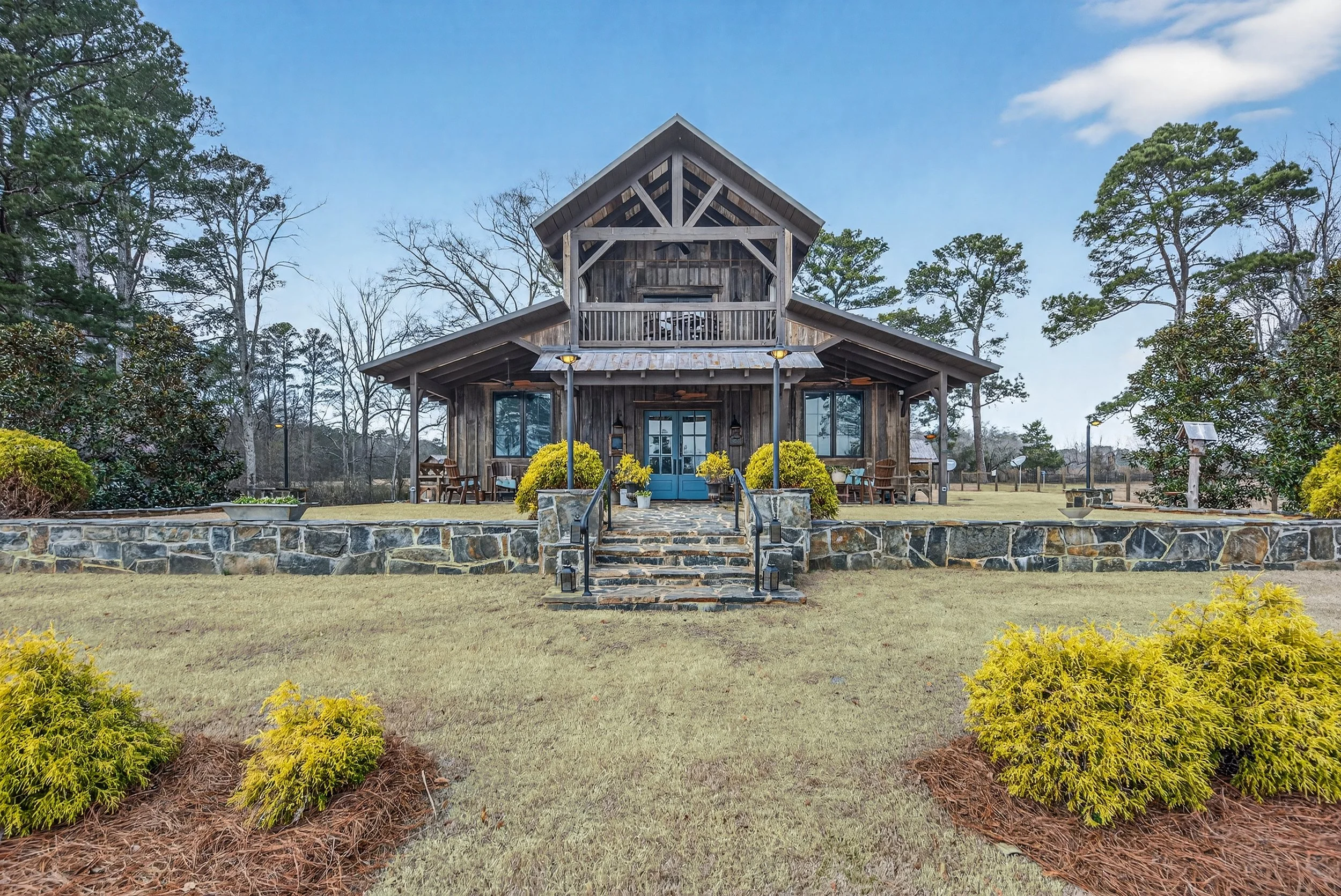 A rustic wooden house with a stone front porch, blue doors, and outdoor seating, surrounded by landscaping and trees under a blue sky.