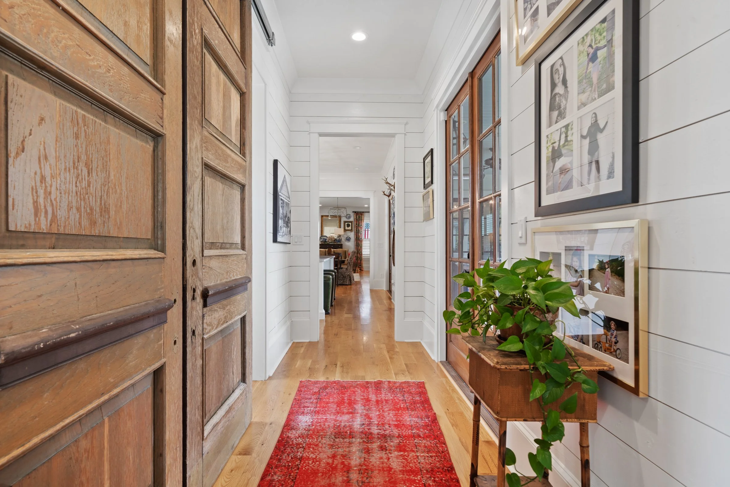 View of a hallway with wooden floors, white shiplap walls, framed photos, a wooden side table with a green potted plant, hallway leading to a living room area with furniture and decor.