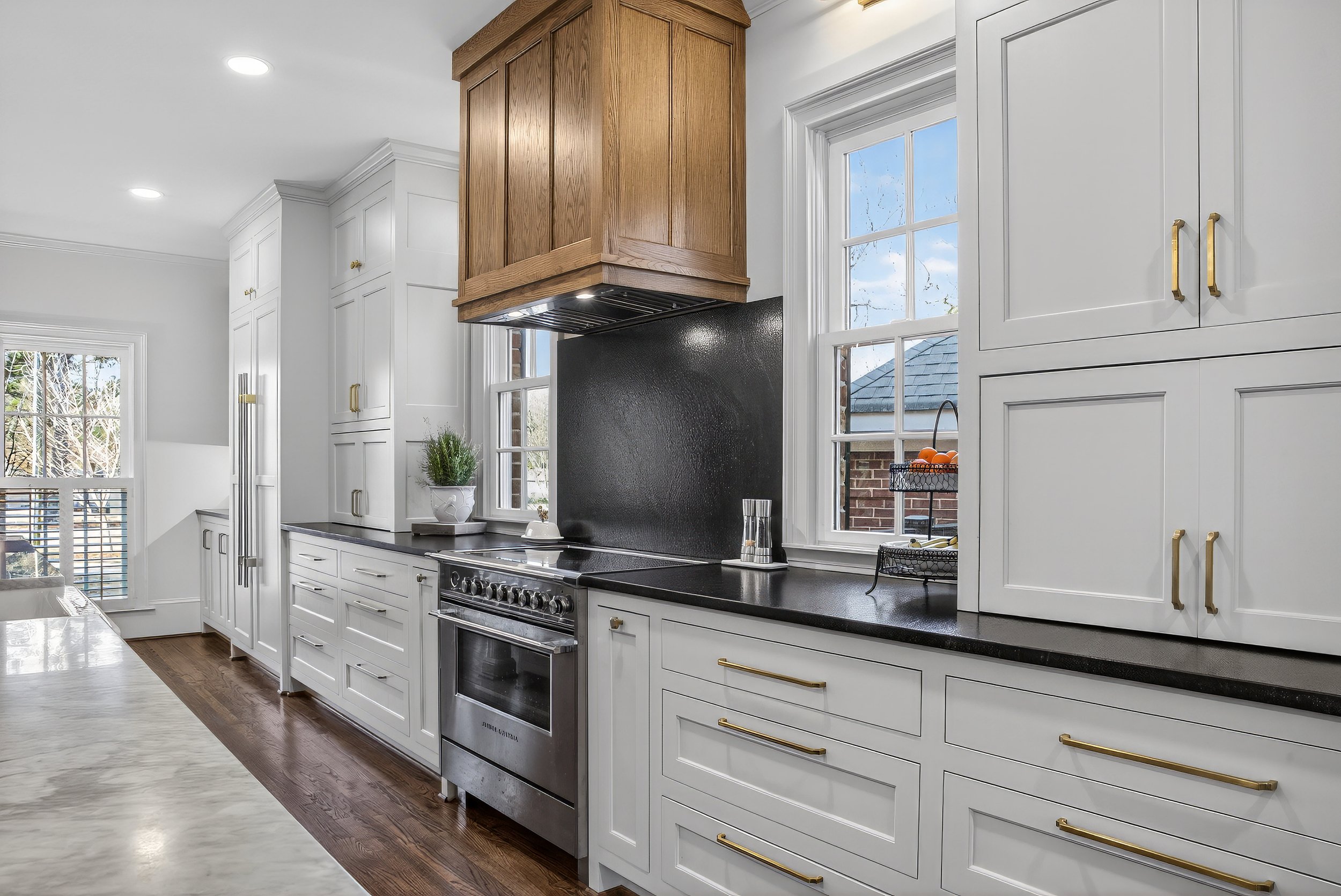 Modern kitchen with white cabinetry, black countertops, a stainless steel oven, and a wooden range hood, with windows showing blue sky and neighborhood rooftops.