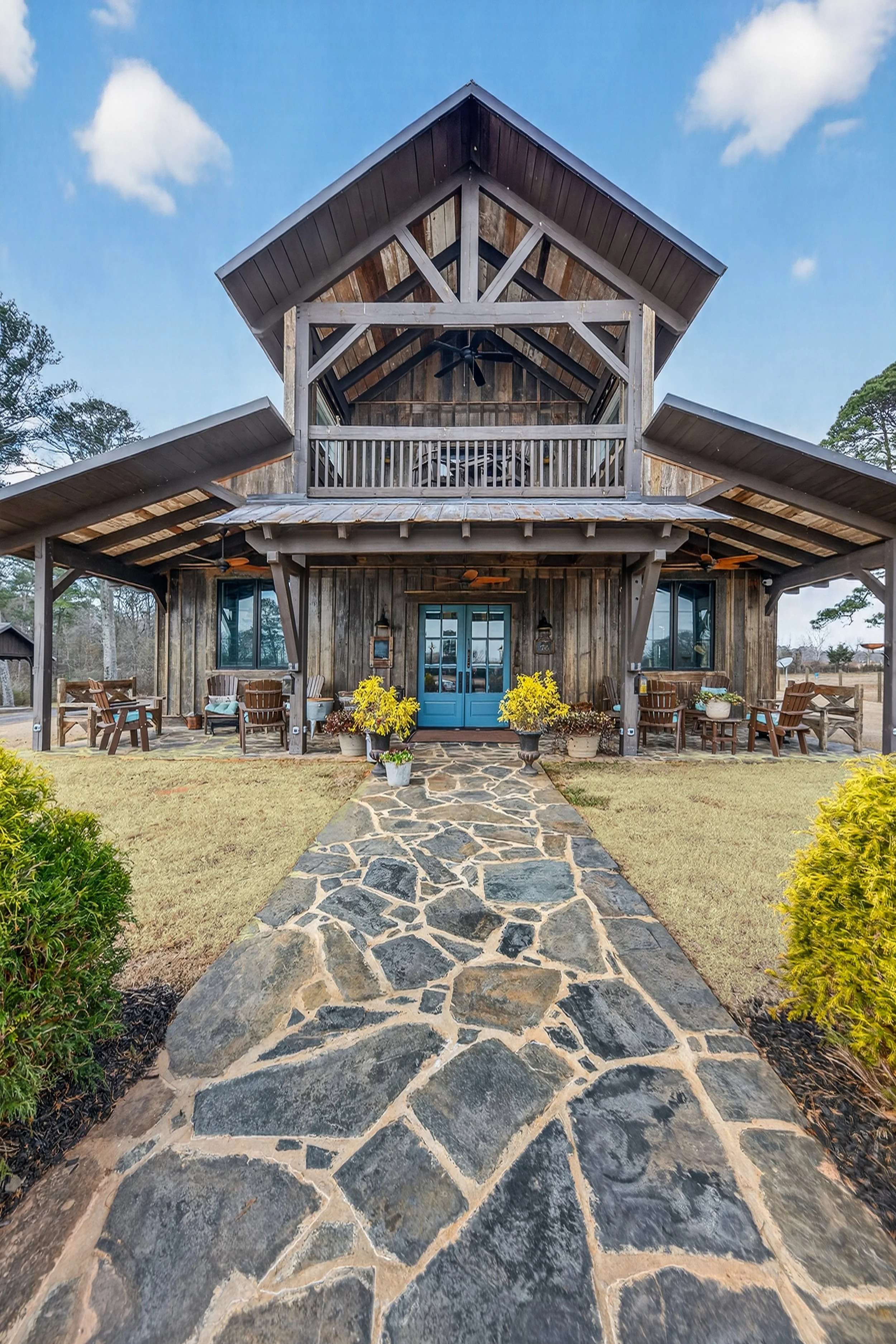 A rustic two-story house with a stone path leading to blue double doors, outdoor seating on a covered porch, and a balcony above, set against a partly cloudy sky.
