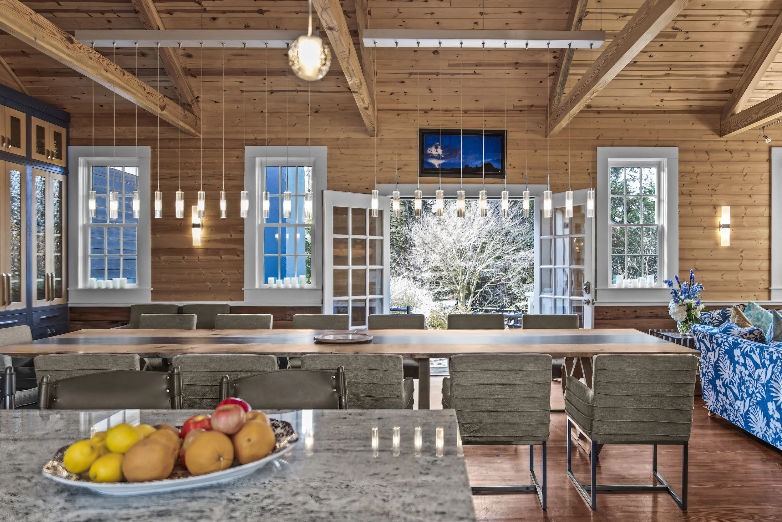 A spacious dining room with a wooden ceiling and walls, large windows, and glass double doors opening to an outdoor view of trees. There is a long wooden table surrounded by padded chairs, a decorative blue floral-patterned sofa, and a plate of fruit