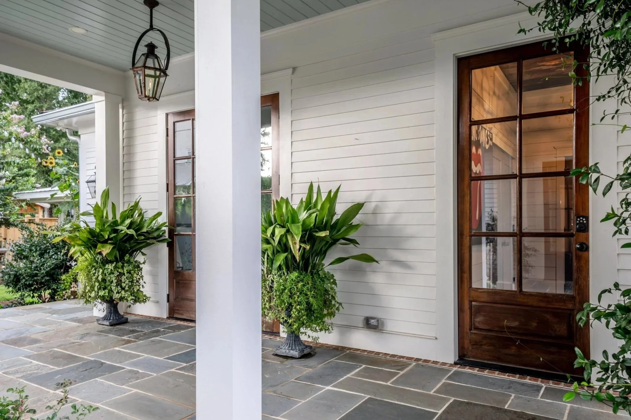 Front porch of a house with potted plants, wooden door with glass panels, white siding, and a lantern hanging from the ceiling.