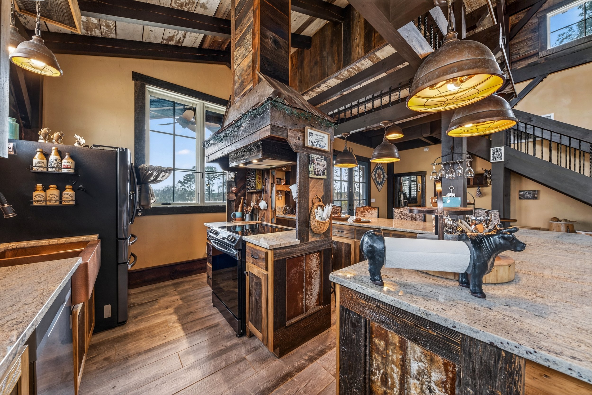 Rustic kitchen with wooden accents, black refrigerator, stove, hanging wine glasses, large island with a decorative black cow paper towel holder, open shelves, and large windows letting in natural light.