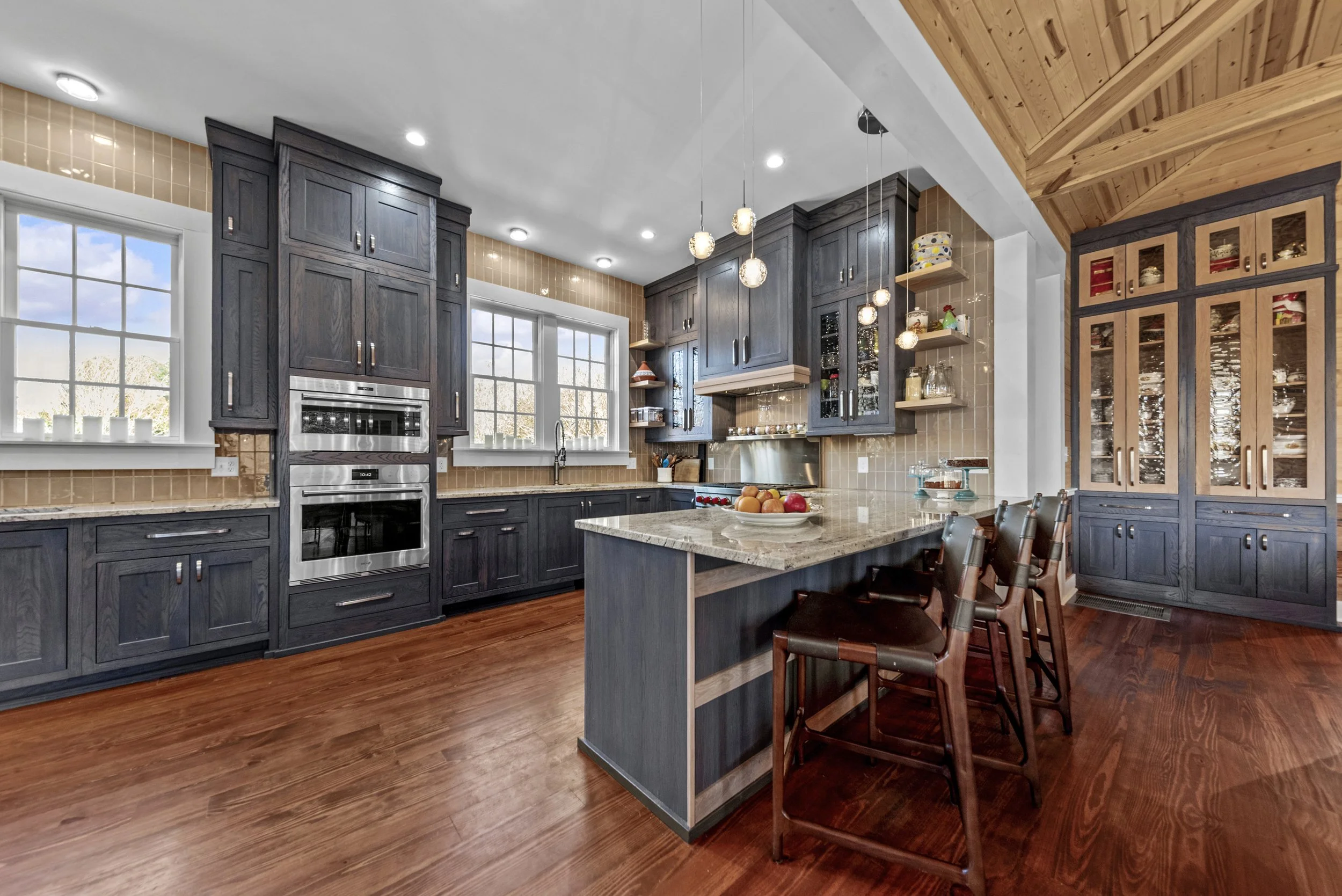 Modern kitchen with dark wooden cabinets, granite island, hardwood flooring, beige tiled backsplash, and natural light from two windows.