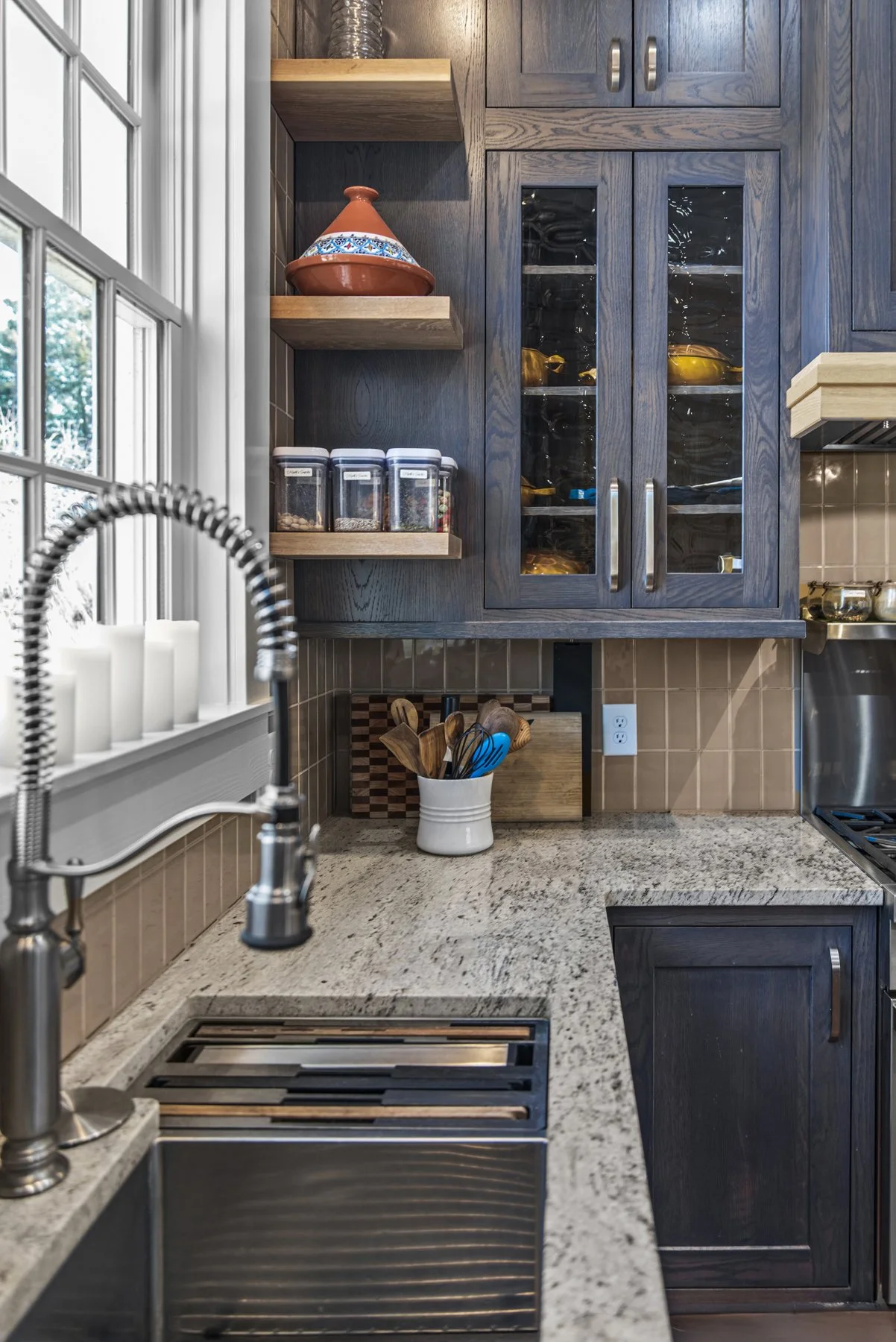 A kitchen countertop with a stainless steel sink, a spring-coiled faucet, a window with white candles, and dark wood cabinets, some with glass doors showing dishes, shelves with spices, and a container holding kitchen utensils.