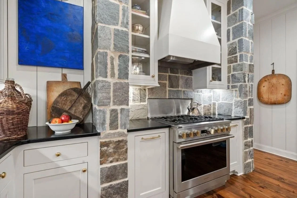 Kitchen with stone walls, white cabinets, stainless steel oven, and wood flooring. Decor includes woven basket, wooden cutting boards, and apples in a bowl.