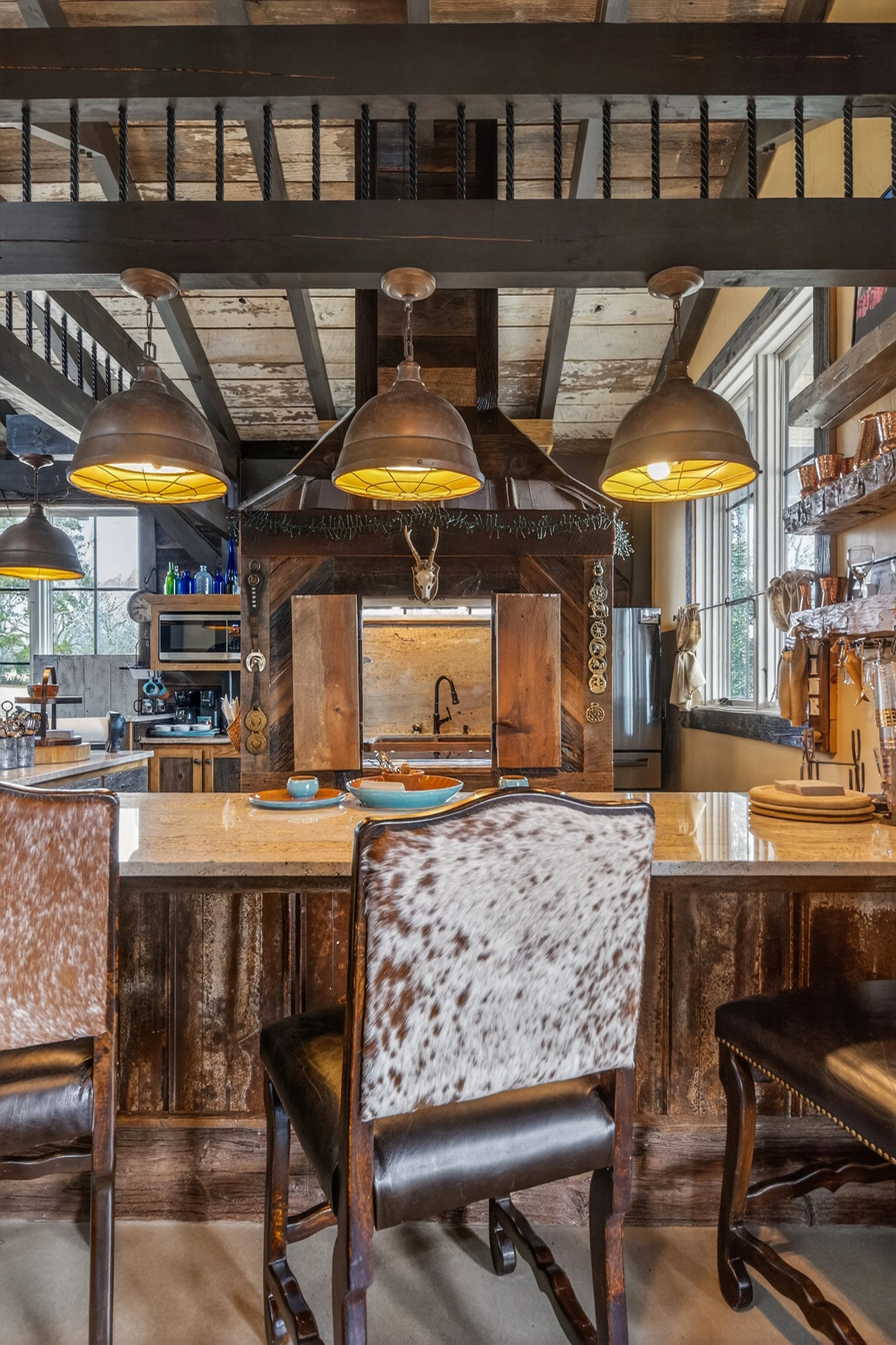 Rustic kitchen with wooden cabinets, cowhide upholstered chairs, hanging industrial-style pendant lights, and a kitchen island with bowls and dishes, large window with natural light