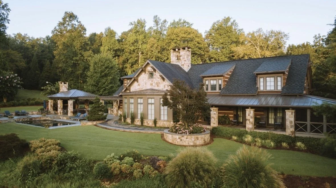 Luxury house with stone exterior, multiple gabled roofs, large windows, surrounded by well maintained lawn and lush trees, with a patio and pool area.