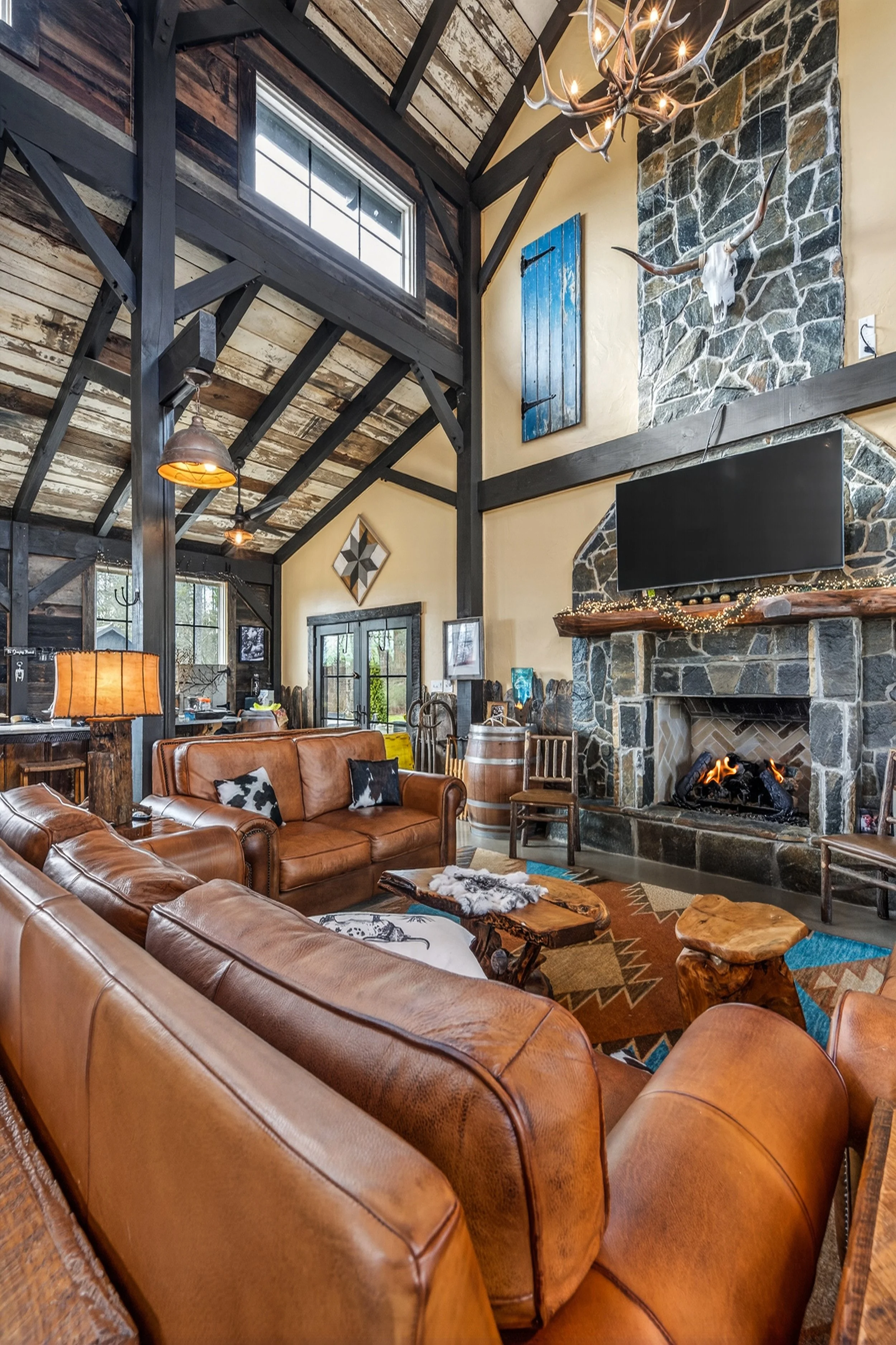 A rustic living room featuring leather sofas, a stone fireplace with a mounted television, a chandelier with antler design, and wooden accents.