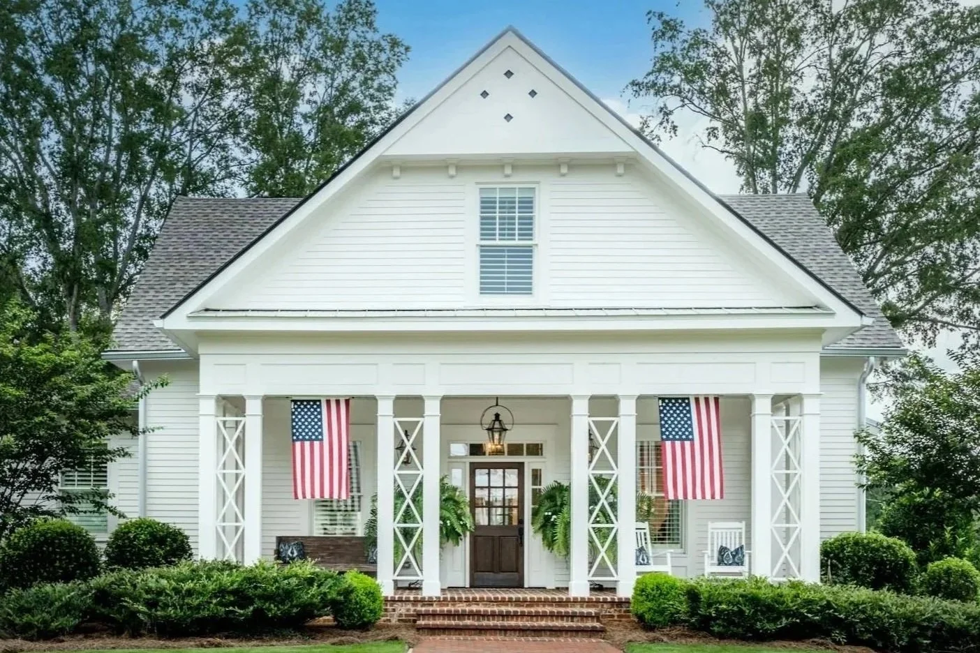 White house with American flags hanging on a porch with green bushes and trees in the background.