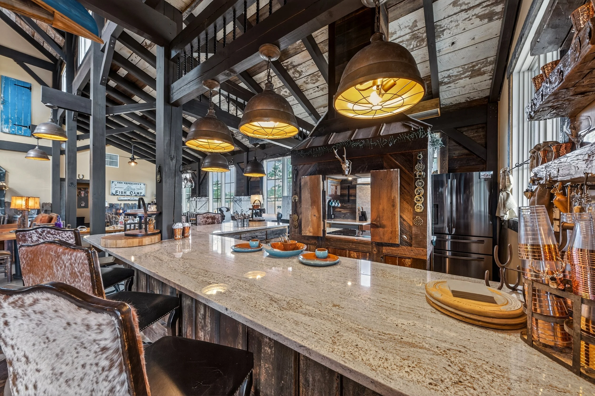 Interior of a rustic-style kitchen or cafe with a large stone countertop, black wooden beams, hanging industrial-style pendant lights, and a variety of dishware and decor.