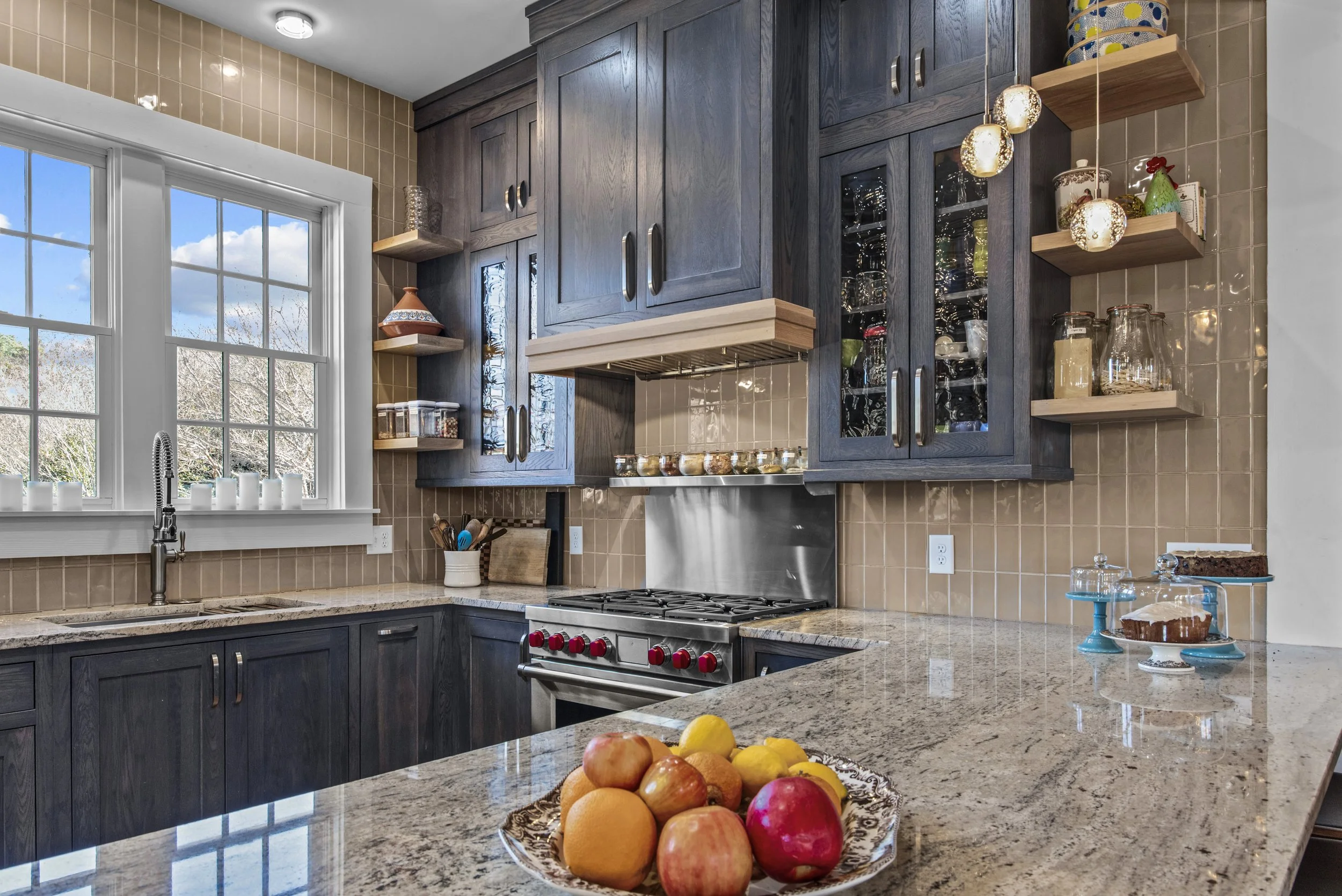Kitchen with dark wood cabinets, beige tile backsplash, granite countertops, a window with candles, and a bowl of apples, oranges, lemons, and a peach on the counter.