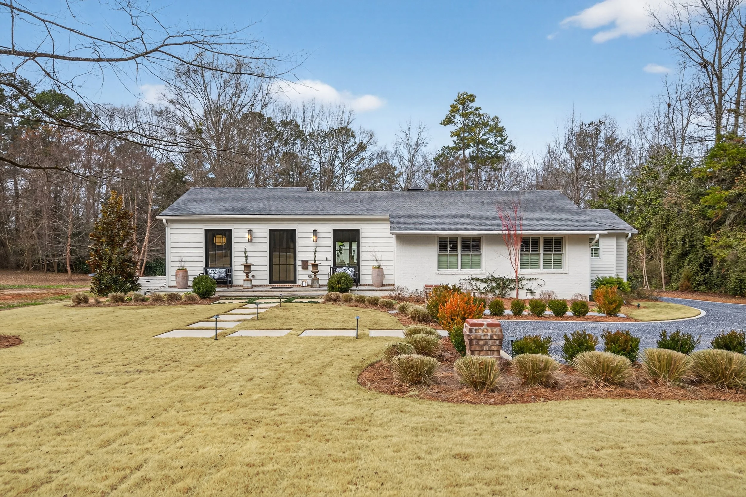 Single-story white house with a gray shingled roof, front porch with seating and decorative planters, large windows with shutters, landscaped front yard with grass, shrubs, and a curved gravel driveway, trees in the background under a partly cloudy sky.