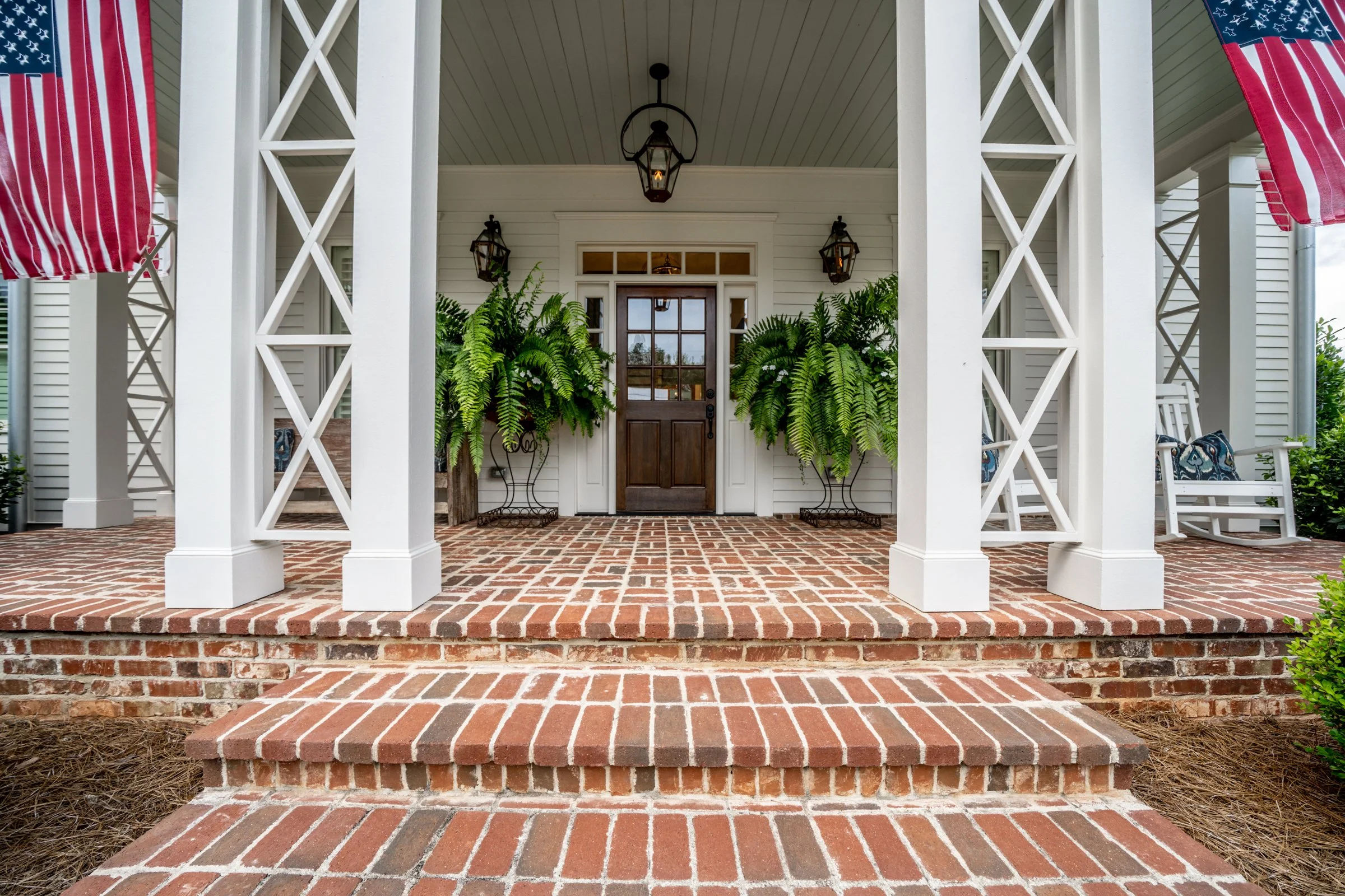 Brick steps leading to a porch with white columns, hanging lantern light, potted ferns, white chairs, and American flags.
