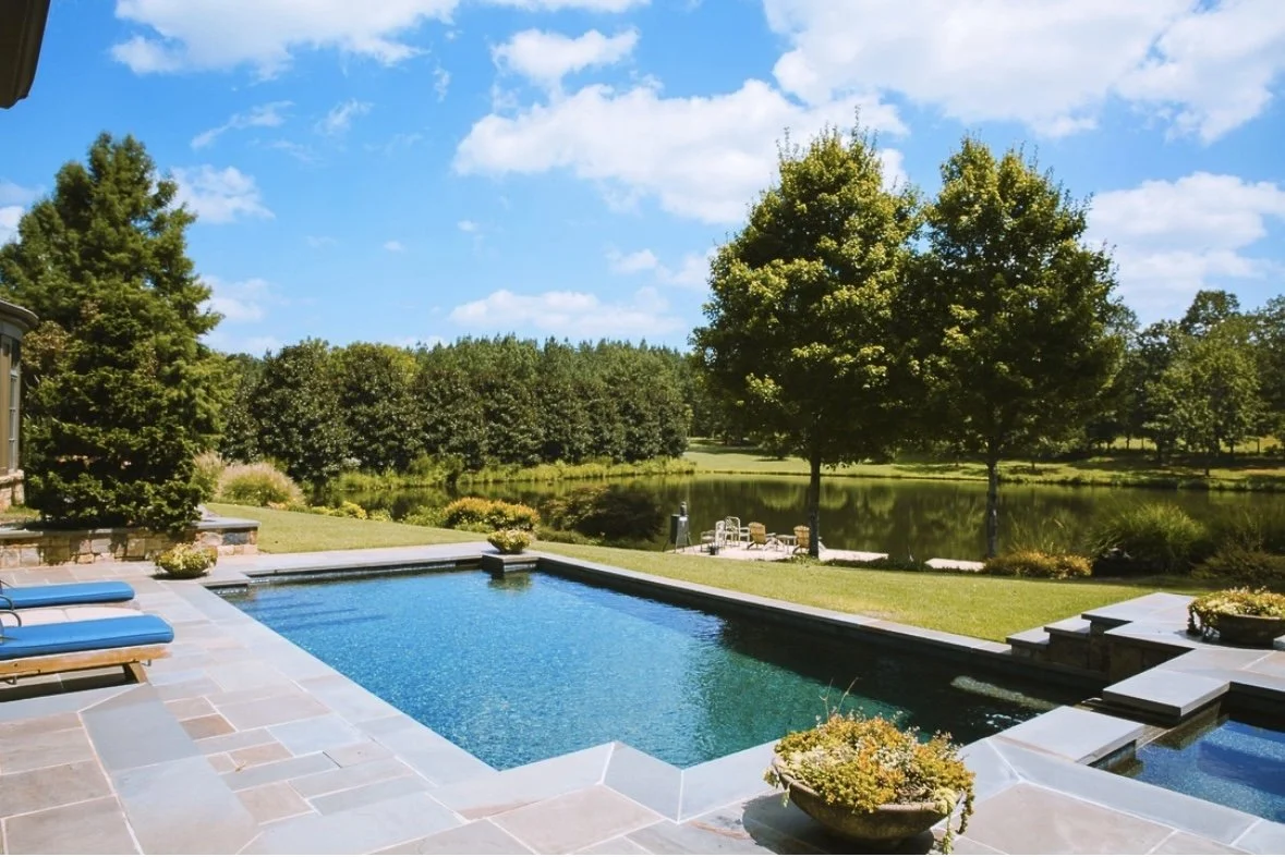 A backyard scene featuring a swimming pool with blue lounge chairs, a grassy area with trees, and a lake in the background under a partly cloudy sky.