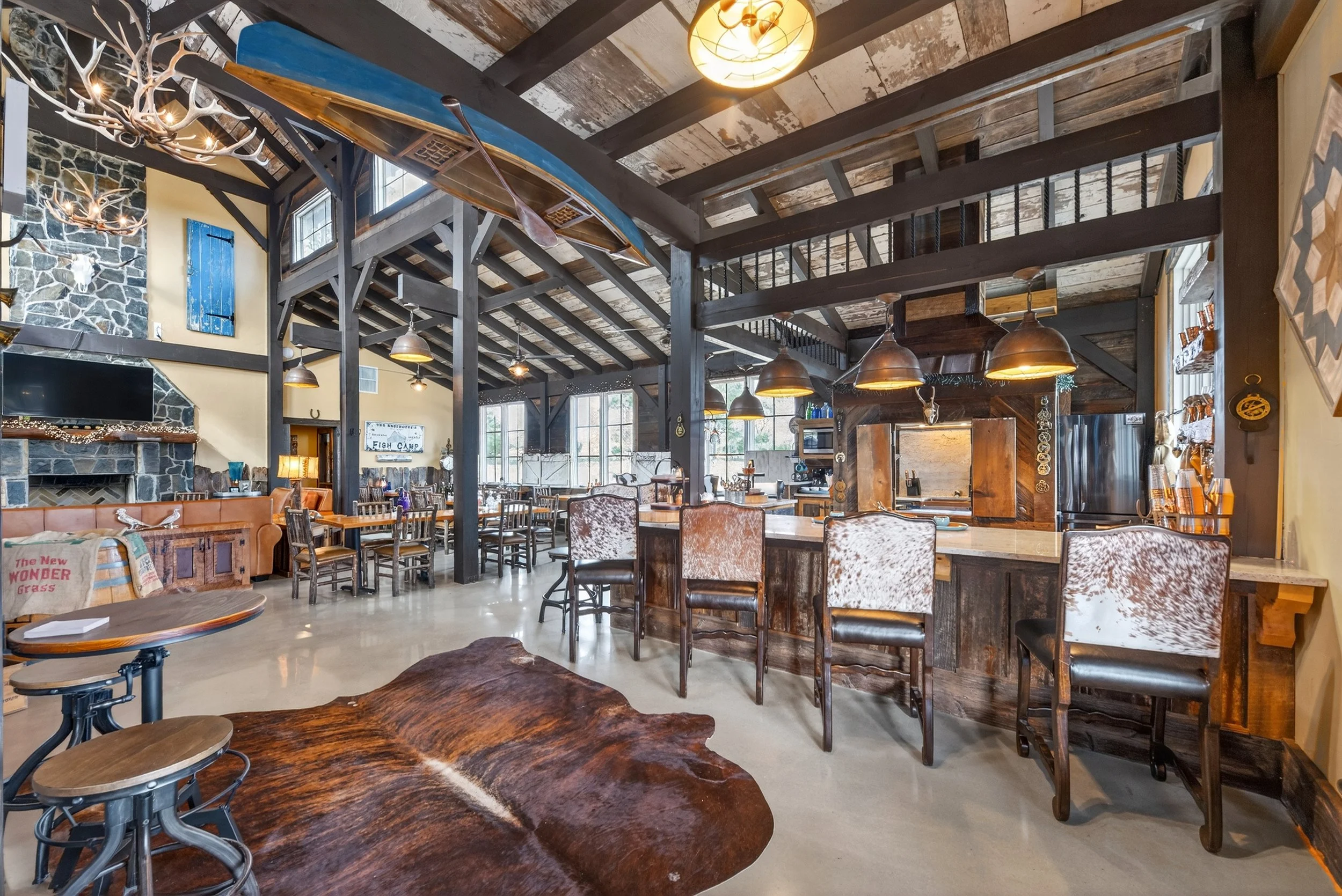 Interior of a rustic restaurant with wooden beams, a bar area with leather and cowhide chairs, large windows, and hanging pendant lights. Decor includes antler chandelier and artwork.