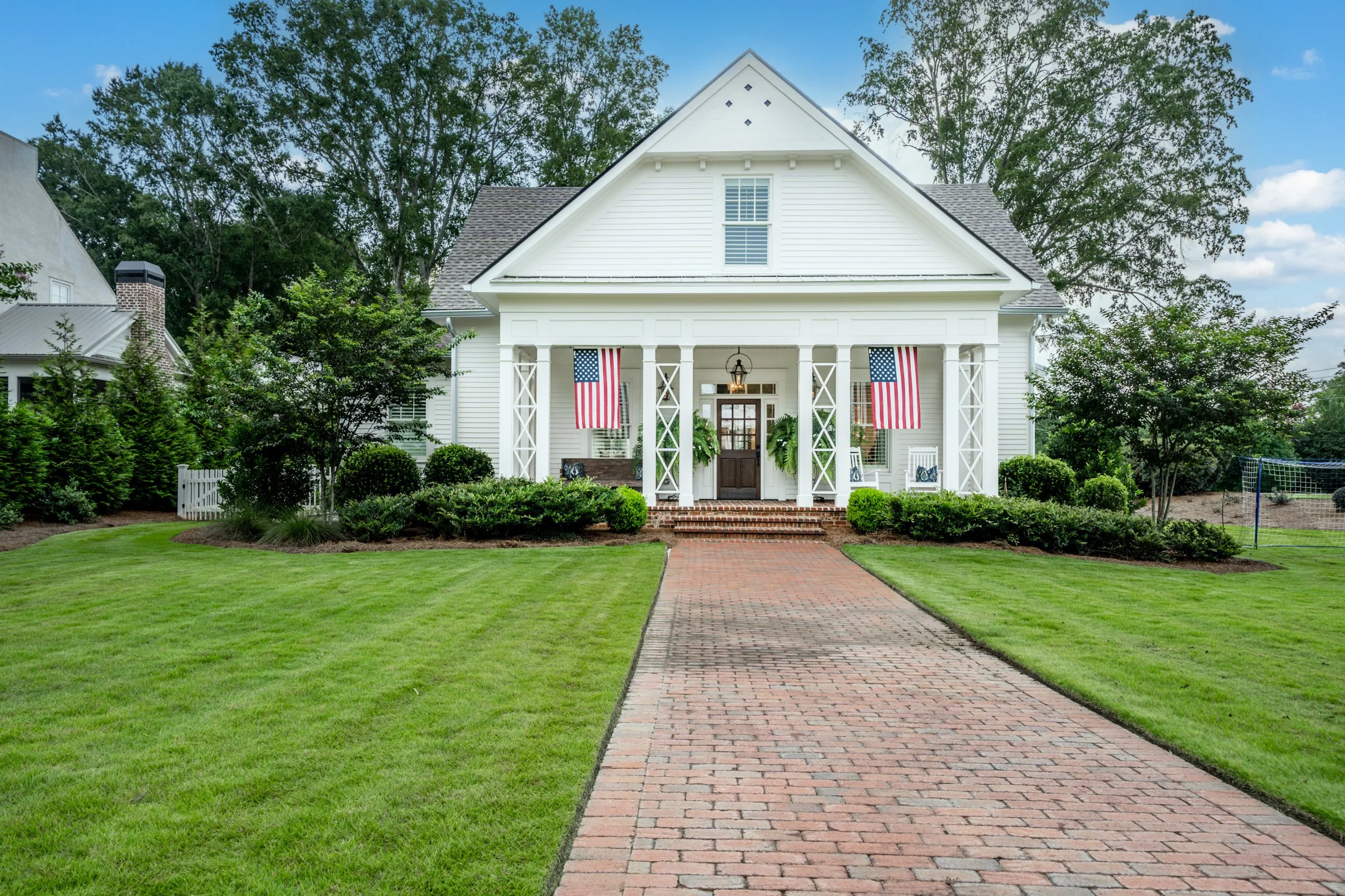 White house with American flags hanging from porch, brick walkway leading to front steps, green lawn and trees in background.