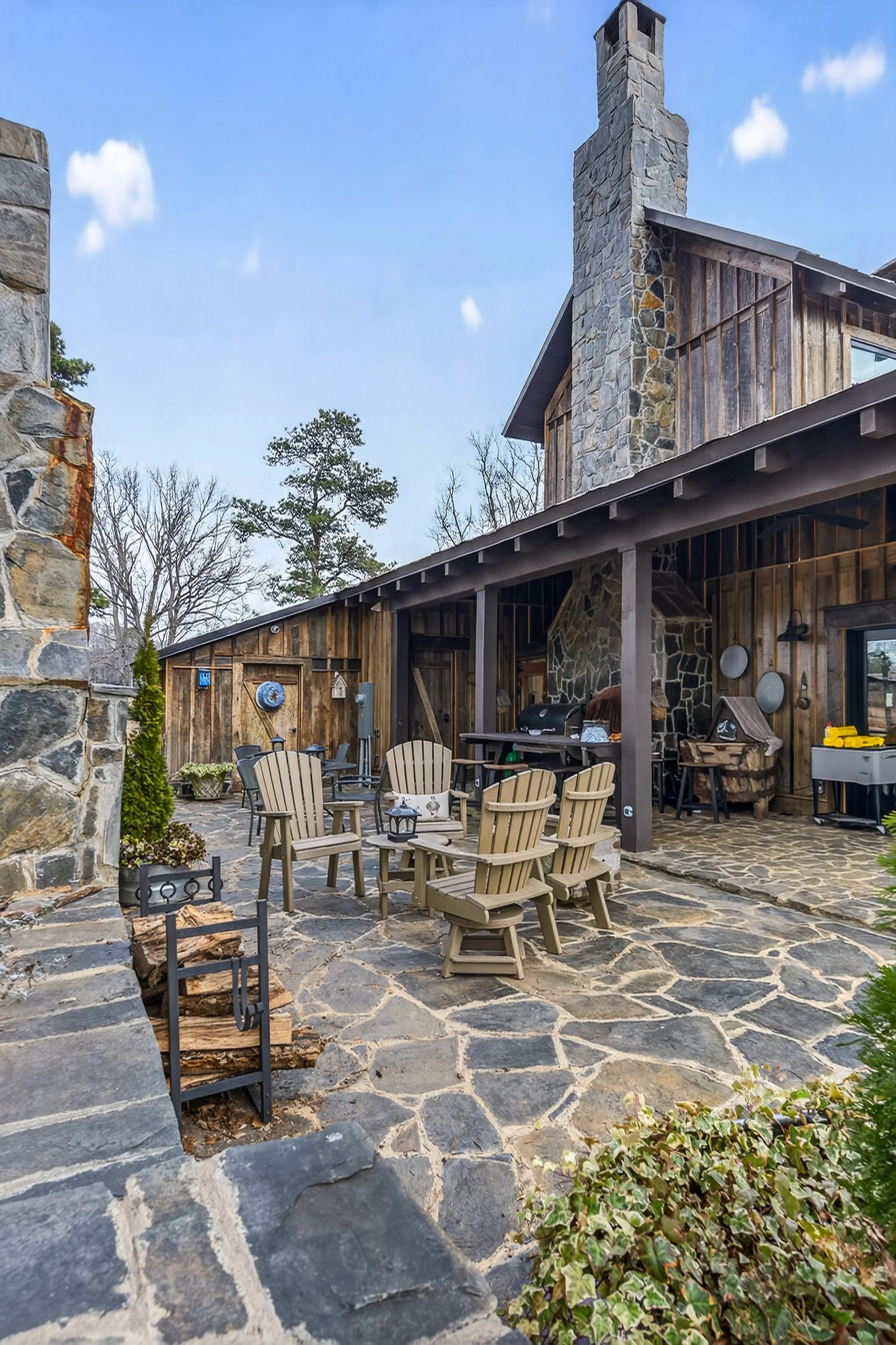 A rustic outdoor patio area with Adirondack chairs, a stone fireplace, and a wooden house with stone accents.