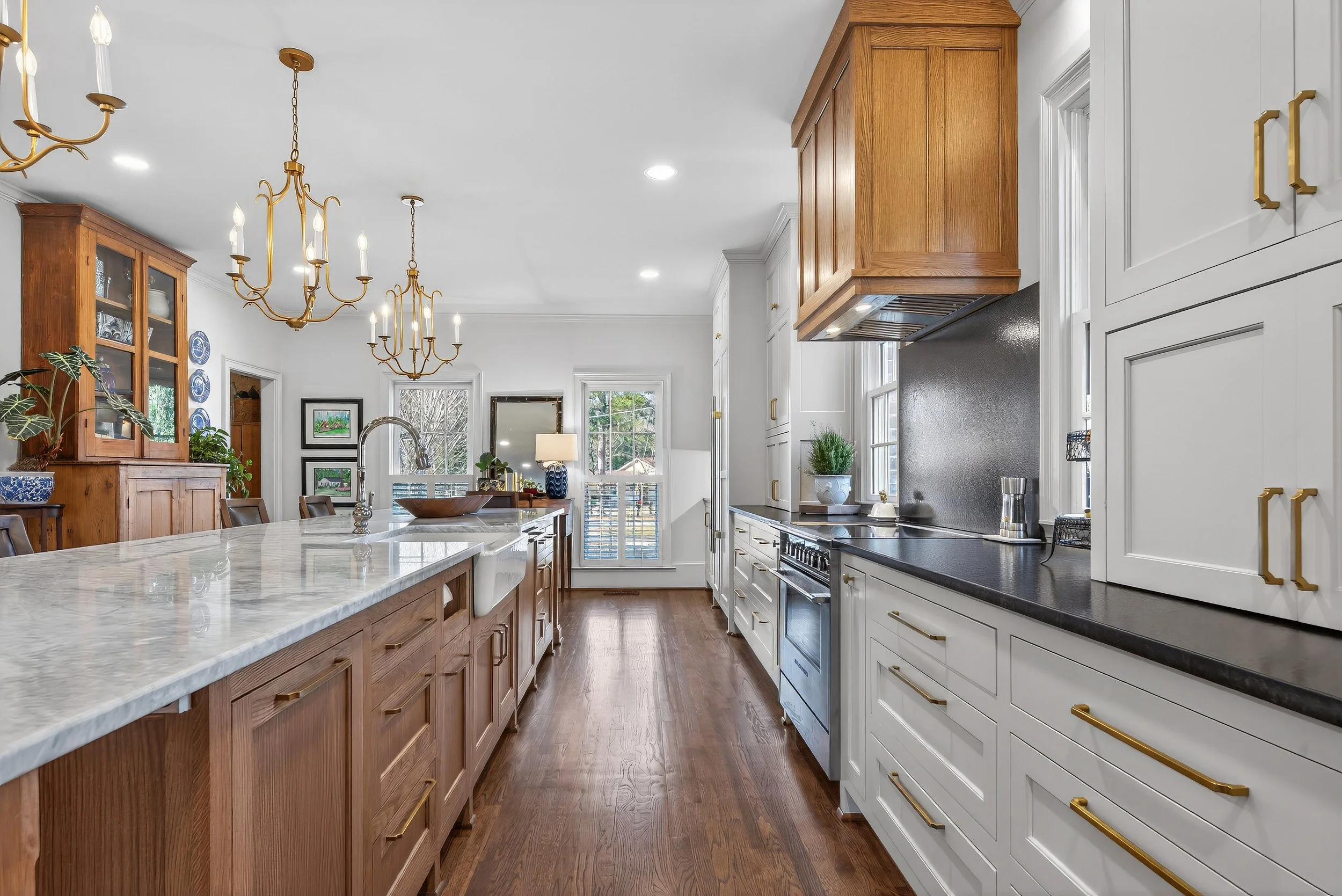 Modern kitchen with white and wooden cabinets, black countertops, and gold hardware, featuring a central kitchen island with a marble top, two gold chandeliers, and a dining area in the background with windows and a door leading outside.