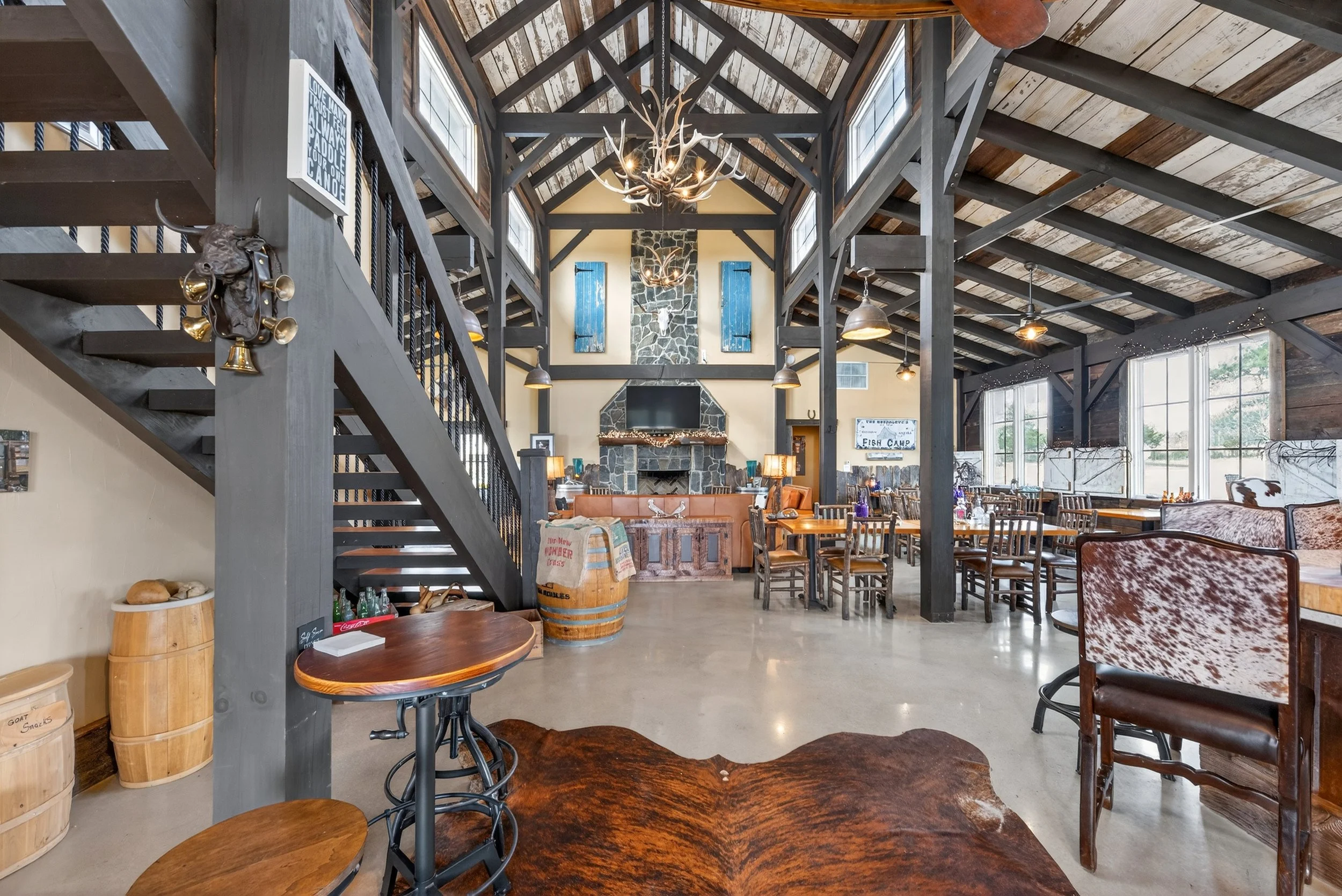 Interior of a rustic lodge or restaurant with a high wooden ceiling, large windows, and a stone fireplace centered on the far wall. The room has wooden tables and chairs, a cowhide rug, and rustic decorations like barrels and antler chandeliers.