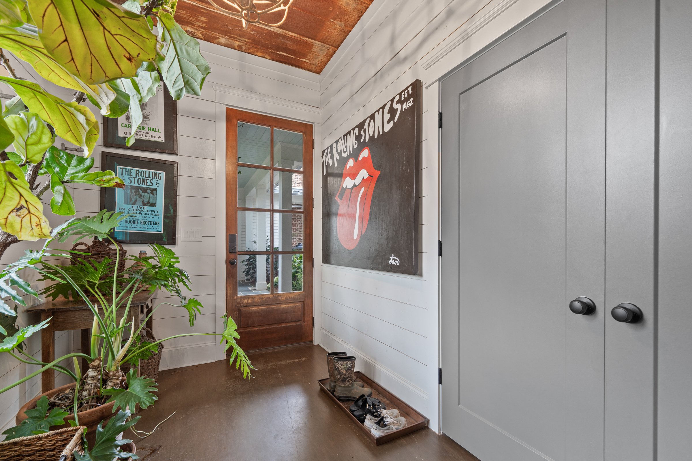 Entryway with a wooden door, potted plants, framed concert posters, a large Rolling Stones artwork with tongue and lips, gray closet doors, and a tray with shoes at the bottom.