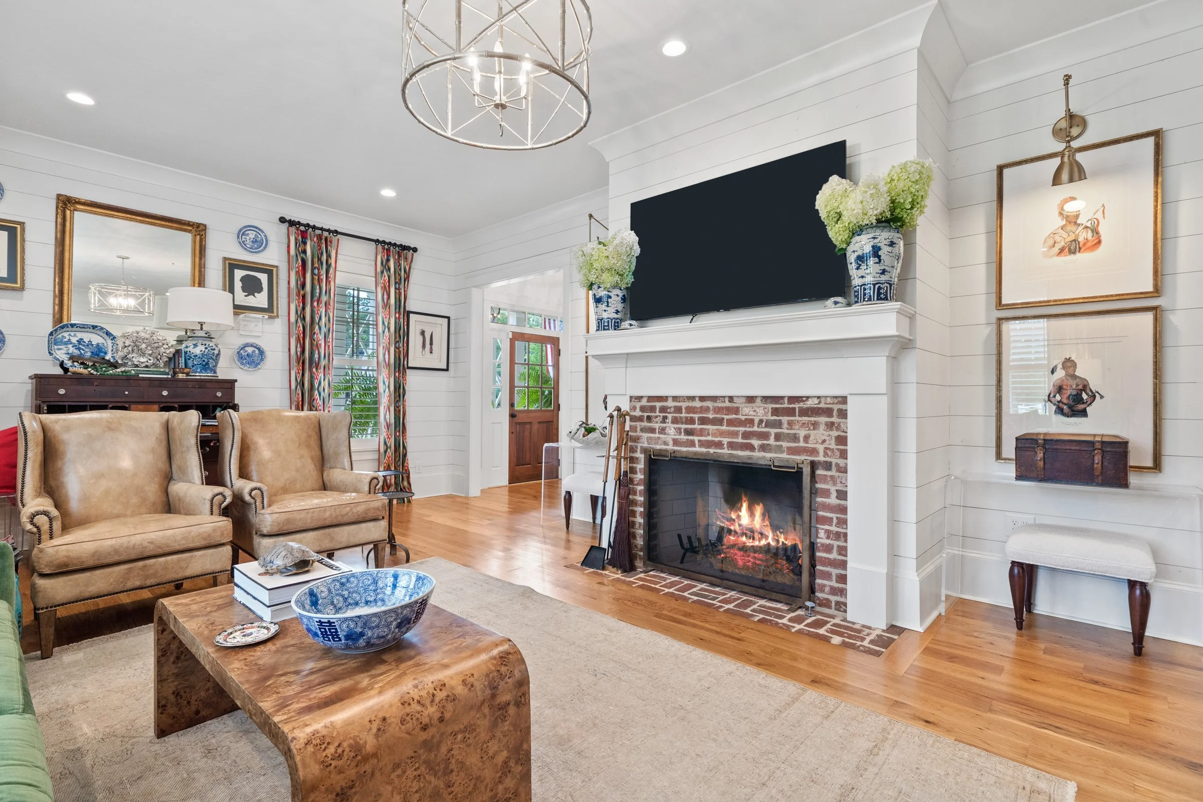 Living room with a brick fireplace, a large flat-screen TV above it, two blue and white vases with flowers on the mantel, a leather armchair, a worn leather couch, a wooden coffee table with decorative bowls and books, colorful patterned curtains, an