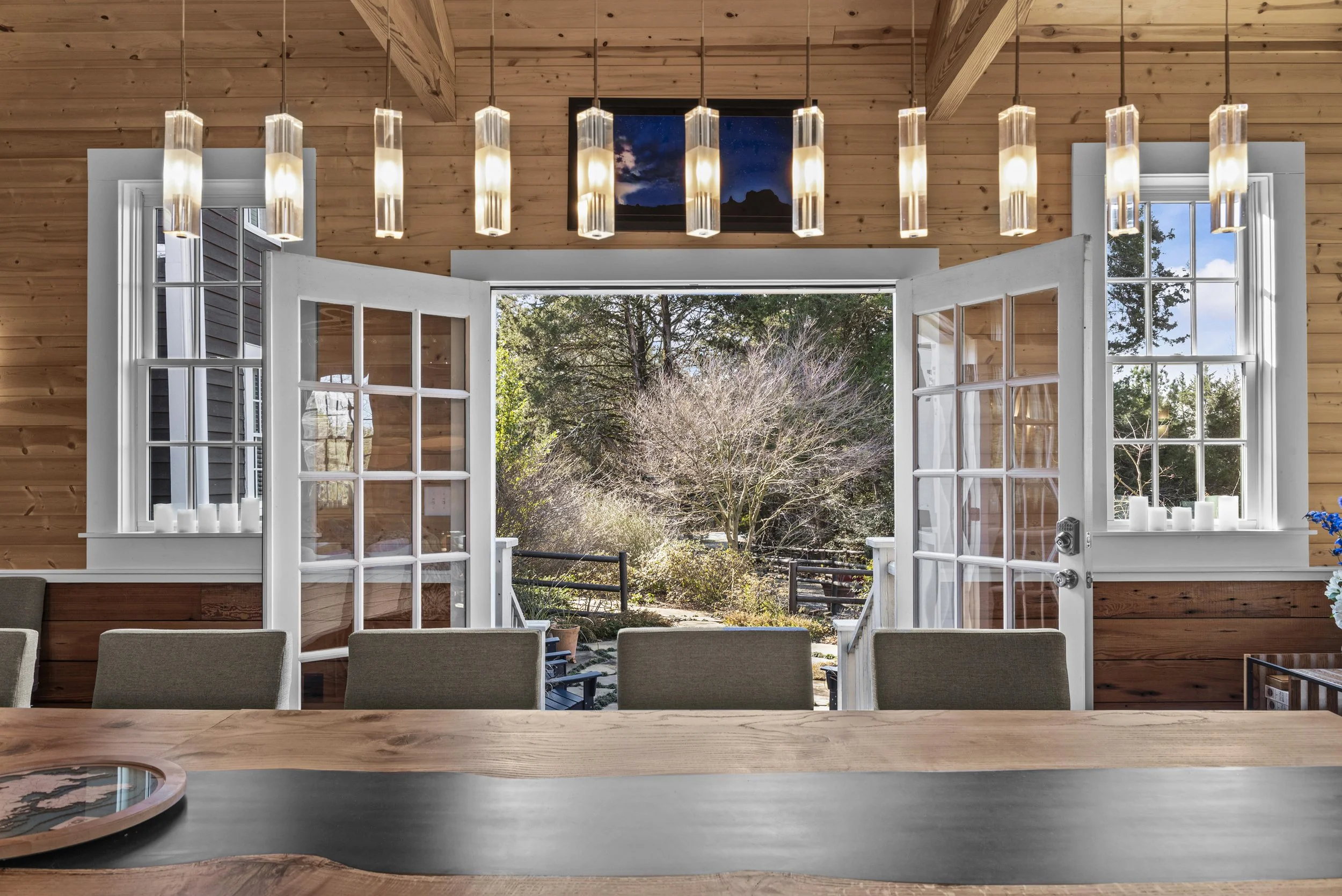 Interior view of a dining room with wooden walls and a large open door leading to an outdoor garden. The room has a wooden table with chairs and modern hanging pendant lights.