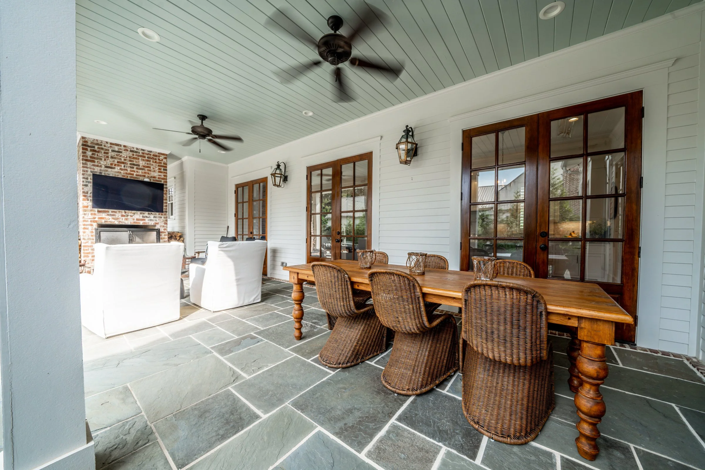Covered patio with wicker dining chairs around a wooden table and white armchairs with a living room featuring a brick fireplace and mounted flat-screen TV in the background.
