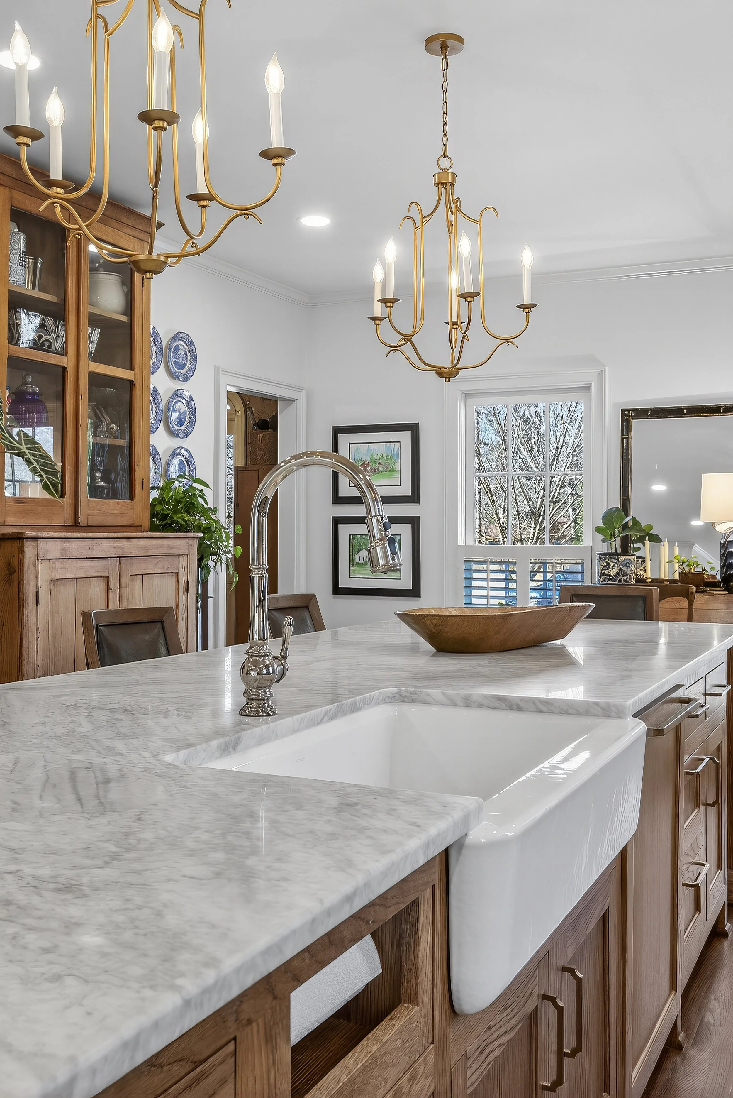 A kitchen with a white marble countertop, wooden cabinetry, a white farmhouse sink, and two gold chandeliers hanging from the ceiling. In the background, there are windows, wall art, and a china cabinet with blue and white plates.