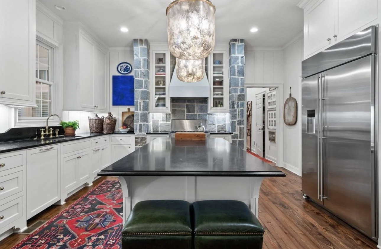 A modern kitchen with white cabinetry, a black countertop, a colorful rug on wooden floors, a large central island with a black surface, a stainless steel refrigerator, and a stone fireplace at the back.
