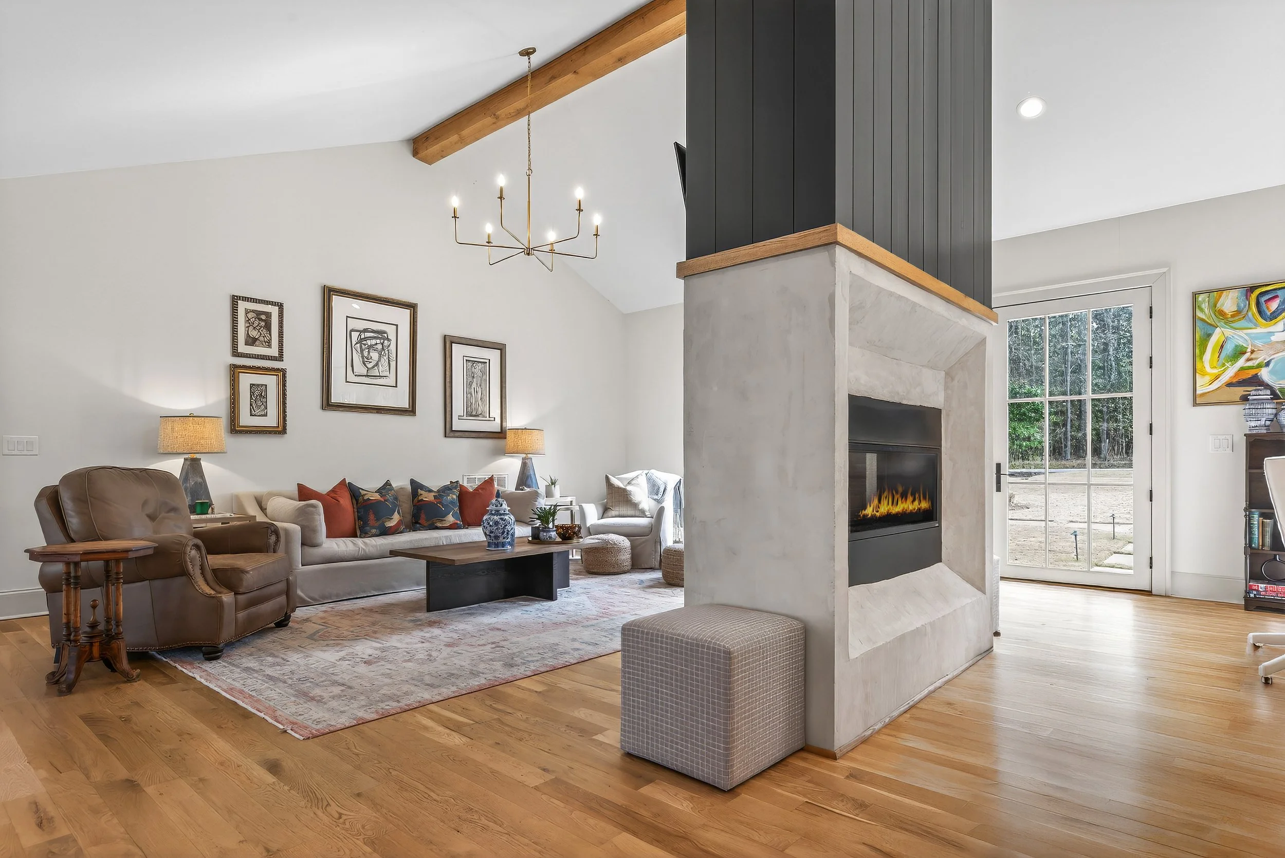 Living room with a fireplace, beige couches with colorful pillows, wooden coffee table, leather armchair, art on the walls, hardwood flooring, and a glass door leading outside.