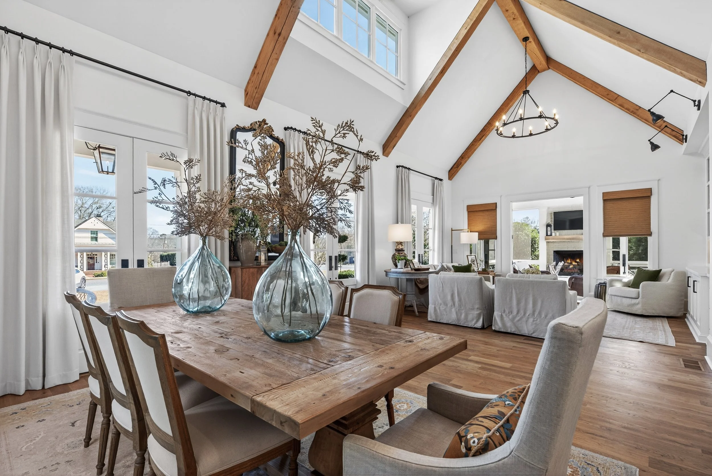 A spacious, bright living and dining room with white walls, exposed wooden beams, large windows, and elegant furnishings. The dining table has two large glass vases with dried branches, and the living area features chairs, sofas, and a fireplace.