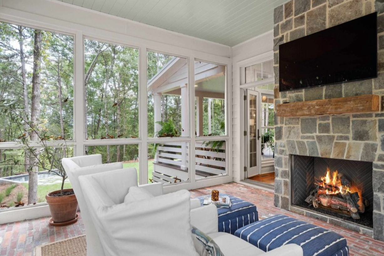 Cozy sunroom with large windows, white armchair with striped blue ottoman, brick floor, stone fireplace with a fireplace and a mounted flat-screen TV, and outdoor trees visible through the windows.