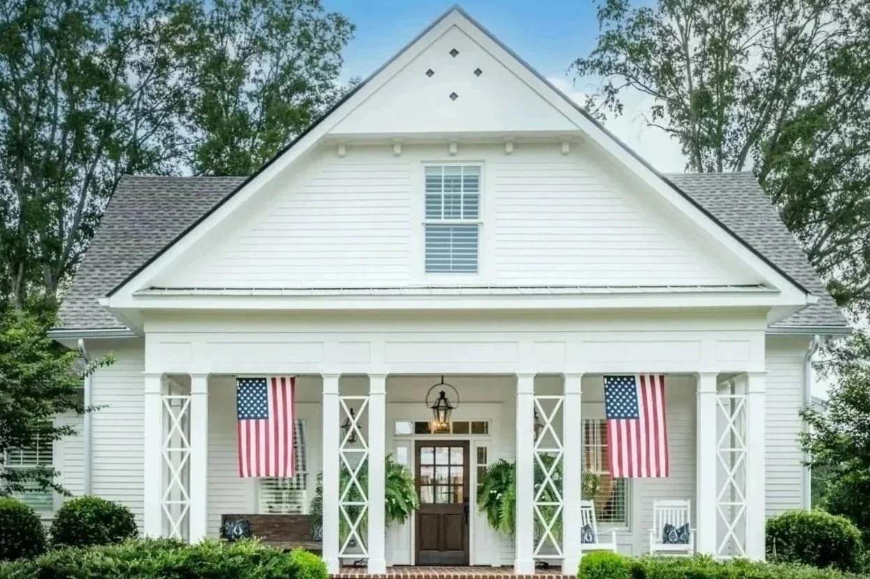 Front view of a white house with a porch, American flags hanging on each side, and a glass front door.