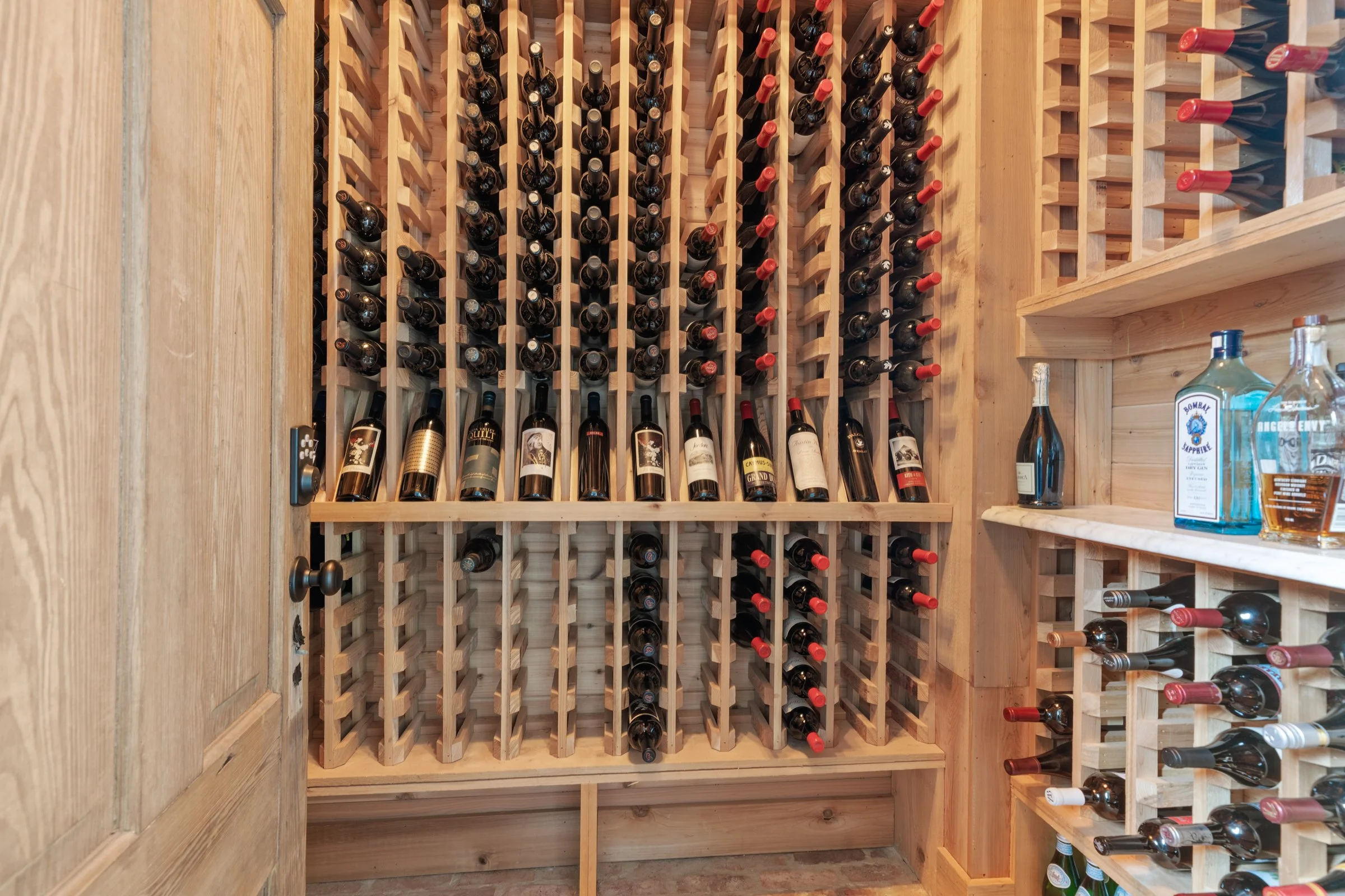 Wooden wine rack with bottles of wine stored horizontally, organized in individual slots with corks showing. Additional bottles are stored on an adjacent wooden shelf.