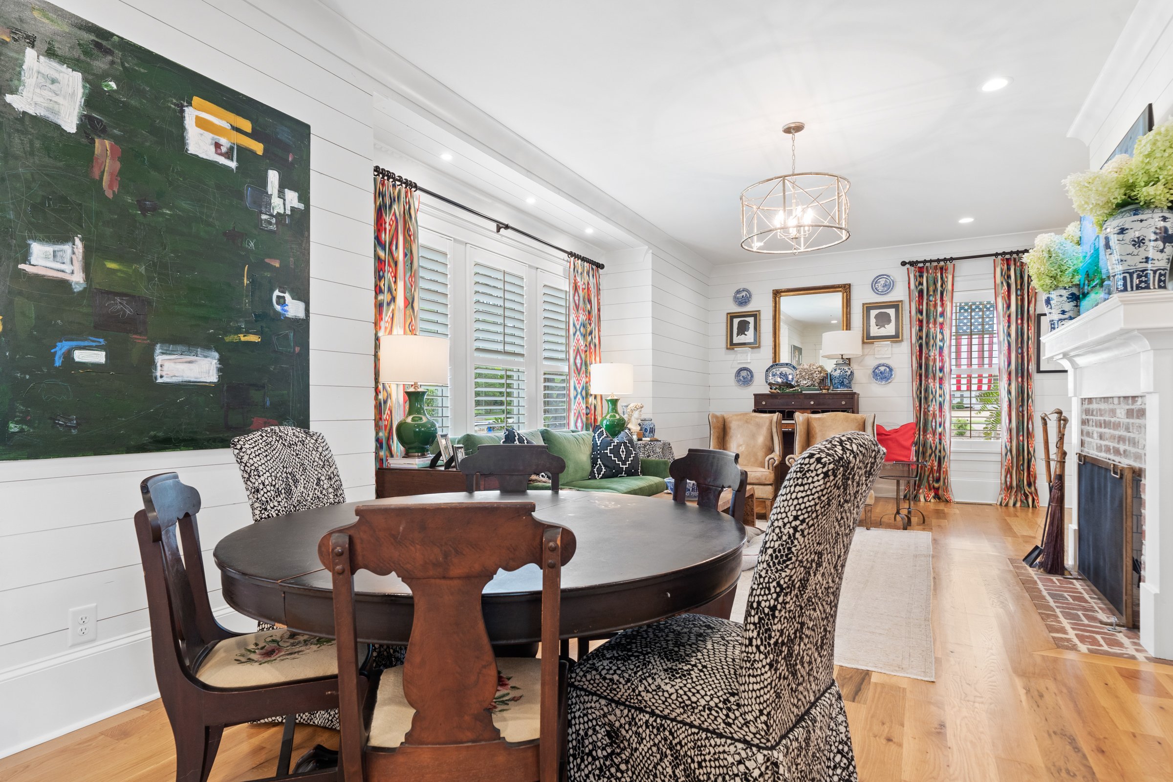 Living room and dining area with wooden flooring, white paneled walls, and colorful curtains. Features a round wooden dining table with mismatched chairs, a green sofa with pillows, and a sitting area with armchairs. Decor includes a large abstract p