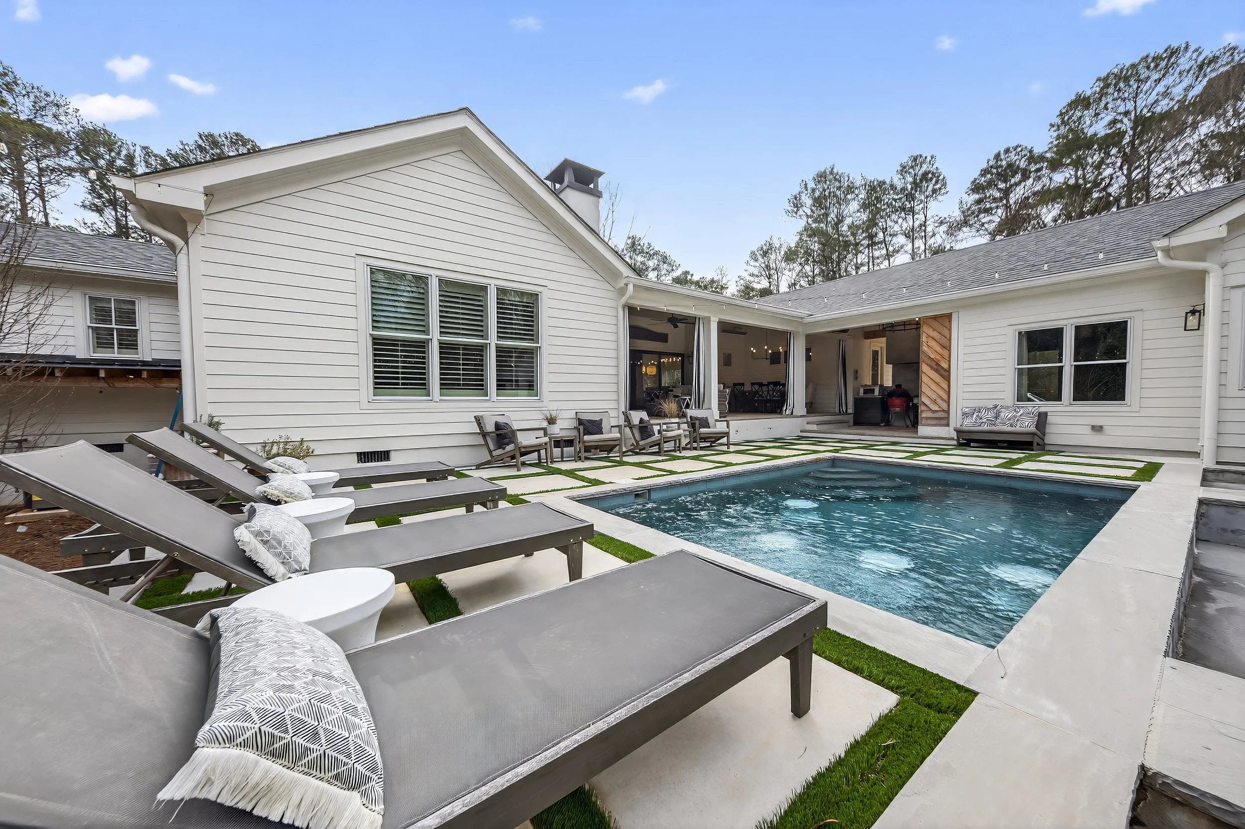 Backyard with a swimming pool, four lounge chairs with pillows, two side tables, outdoor chairs, a bench, and a house with large windows, a covered patio, and a door.