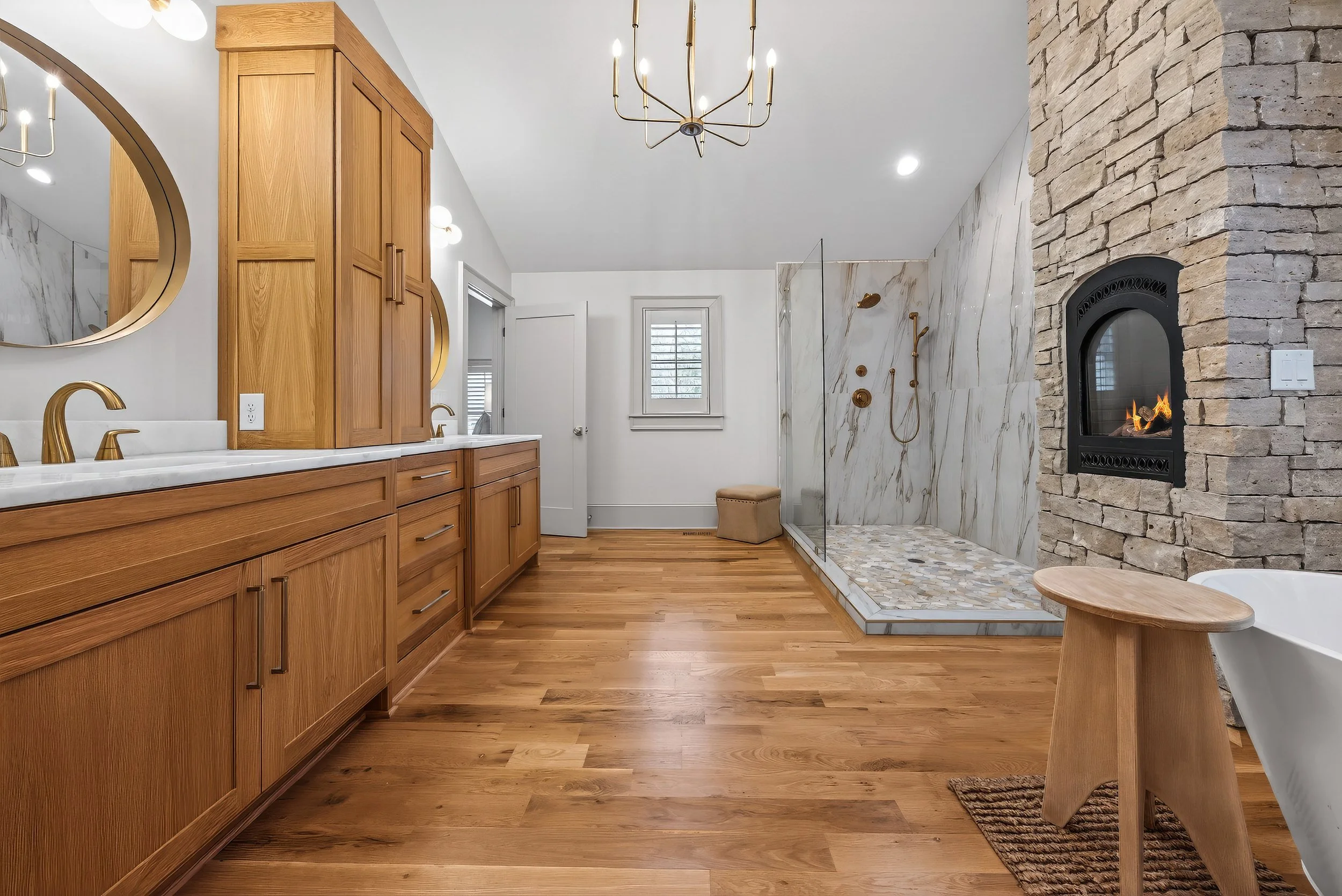 Bathroom with wood cabinets, marble countertops, gold fixtures, a shower with marble and pebble tile, a stone fireplace with a fire, a white freestanding tub, and hardwood floors.